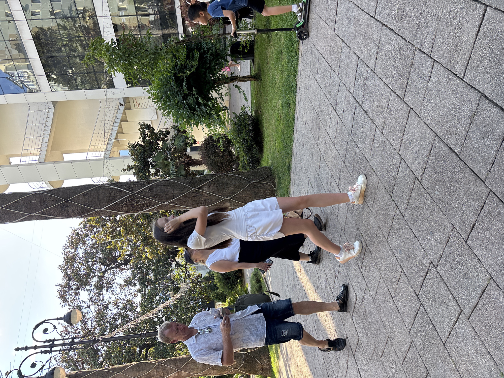 The image captures a sunny day in Sochi, Russia, with several people in a park-like setting. The primary subjects are a man, a woman, and two children.
The man, with blond hair, is wearing a light-colored, short-sleeved patterned shirt and dark denim shorts. He is holding a mobile phone in his hand and appears to be looking at it. He is wearing dark sandals.
A woman, wearing a white dress and white sneakers, is in the foreground. She is leaning back and appears to be interacting with a younger person. Her arm is bent behind her head.
Behind the woman, a child is seen wearing a white t-shirt and dark shorts, also in white sneakers. Further in the background, another child is riding a scooter. This child is wearing a blue t-shirt and black shorts and is on a black and green scooter.
The scene is set on a paved walkway made of large, grey rectangular tiles. To the left of the walkway is a patch of green grass, with trees and lush foliage. In the far background, a modern building with a reflective glass facade is visible. The overall impression is a casual outdoor scene on a bright, likely warm day, given the attire of the people. The image orientation is portrait, with the subjects mostly facing towards the right of the frame. The time of day appears to be daytime, with clear lighting.