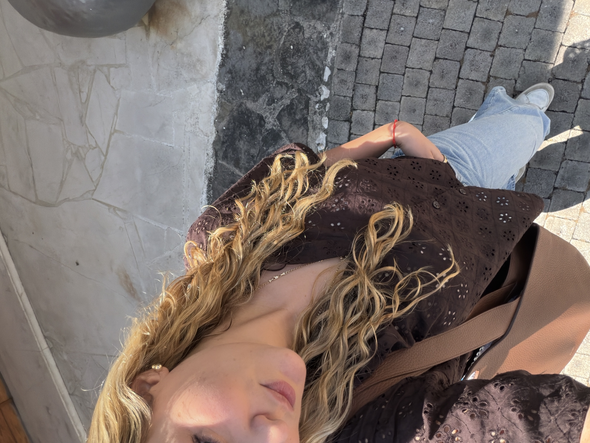 A woman with long, curly blonde hair is wearing a dark brown, eyelet-patterned shirt and light-blue jeans, standing on a gray cobblestone surface. A tan-colored purse or tote bag is slung over her shoulder. The background includes a gray, textured stone wall or building on the left, and a more weathered gray surface that transitions to the cobblestones. The photo appears to be taken from a high angle, looking down at the woman. There is no confirmation of the location being San Bernardino Tlaxcalancingo, Mexico.