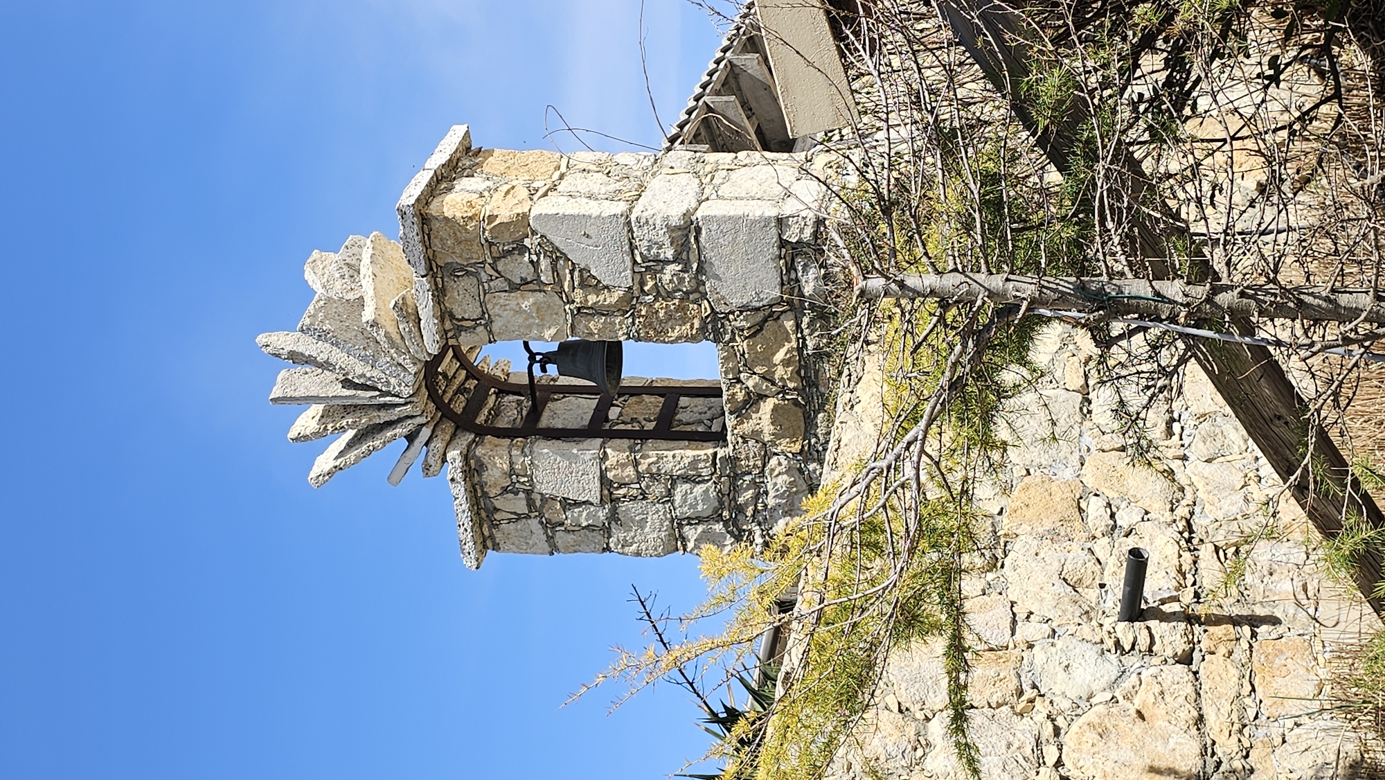 This outdoor image captures a rustic stone structure, likely a small bell tower, set against a vibrant clear blue sky in Varna, Bulgaria. The main subject is a sturdy bell tower constructed from irregular, light-colored stone blocks, giving it an aged and robust appearance. An arched opening in the tower frames a dark, metallic bell suspended from an iron framework. Above this opening, a distinctive decorative feature is visible, formed by several flat, rectangular stones fanning out in a radial or star-like pattern.
To the right of the bell tower, a textured stone wall continues the architectural theme, partially obscured by a dense arrangement of tree branches. These branches, some thick and gnarled, others slender and intricate, are mostly bare, but some sections show sparse green, needle-like foliage, suggesting an evergreen species. A wooden beam or rafter is visible in the upper right background, integrated with the building structure. Towards the bottom right of the frame, a short, dark pipe protrudes from the stone wall.
The scene is bathed in bright daylight, indicated by the clear blue sky and the crisp shadows cast by the branches and architectural elements, highlighting the texture of the stone and the intricate network of the tree limbs. The overall impression is one of historical charm and natural integration, with no people or visible text present.