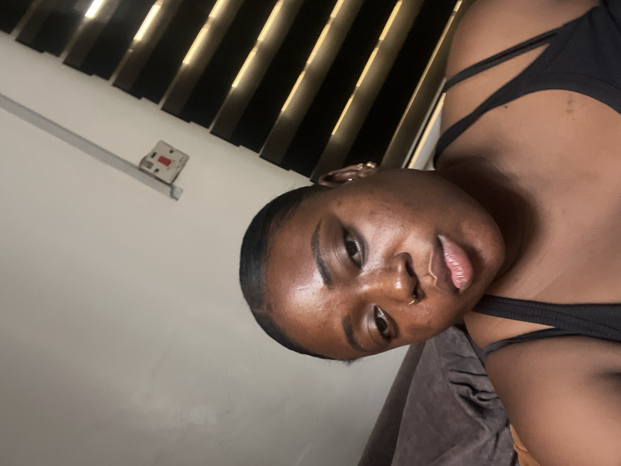 A close-up, slightly angled shot shows a dark-skinned woman with short, slicked-back hair. She has a nose ring in her left nostril and earrings. She's wearing a black top with multiple straps on the shoulder. The background includes a wall, blinds with light filtering through, and a switch on the wall. The location and city are unknown.