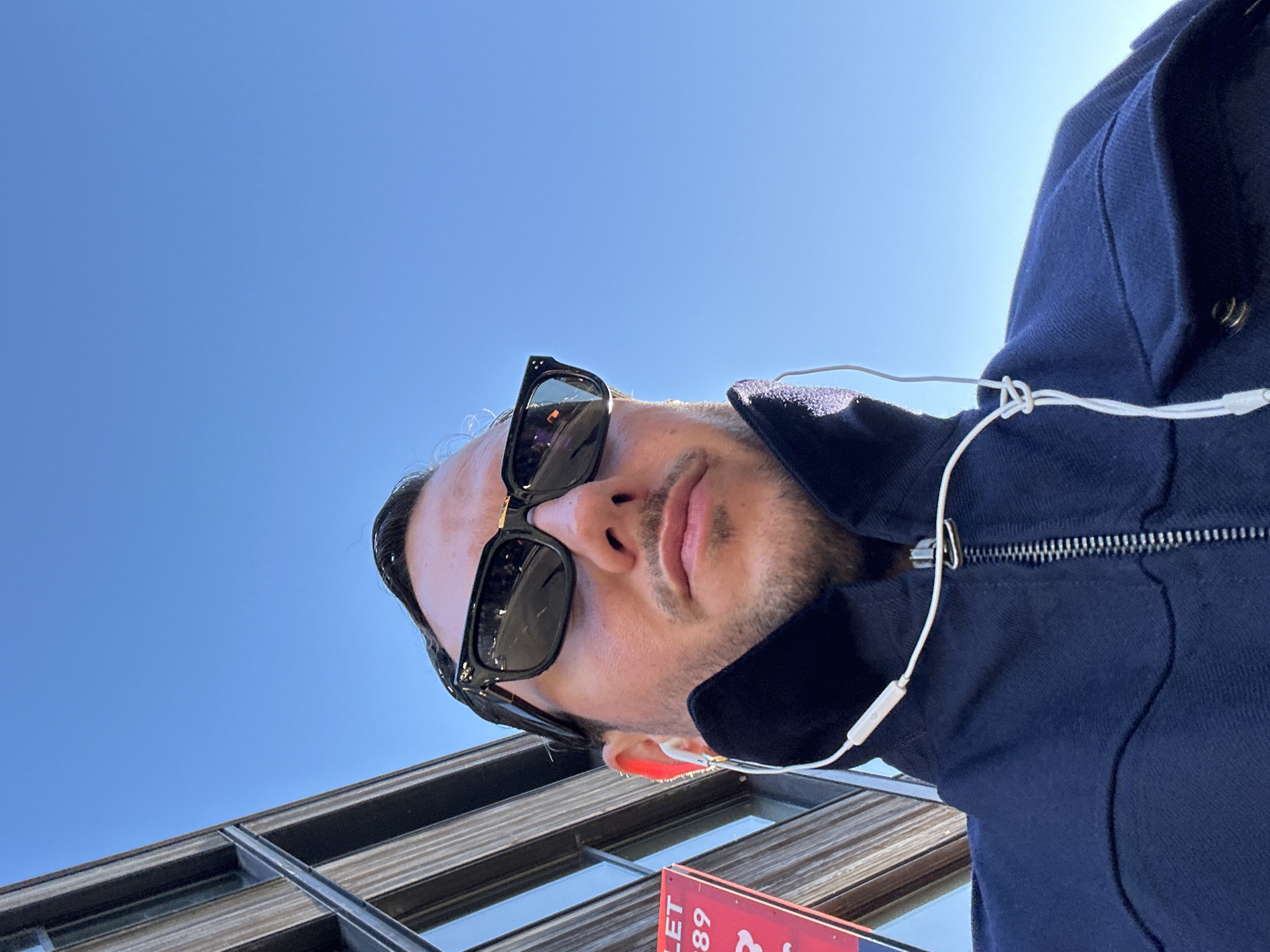 A low-angle, close-up shot captures a man from the forehead to the mid-chest, looking directly towards the camera, likely in a selfie-style pose. He is wearing dark rectangular sunglasses, a dark blue zip-up jacket or hoodie, and white wired earphones, one of which is visible in his right ear. His facial hair includes a short beard and mustache.
The background is predominantly a clear, bright blue sky, indicating a sunny day. Below and to the left of the man, a modern building structure with dark, horizontal frames and reflective surfaces is partially visible. In the bottom right corner, a red sign with fragmented white text is seen. The visible text includes "...LET...", "489...", and a "£" symbol, strongly suggesting a property "To Let" sign with a price. The setting appears to be an urban environment, consistent with the provided London context. The overall lighting is bright and natural.