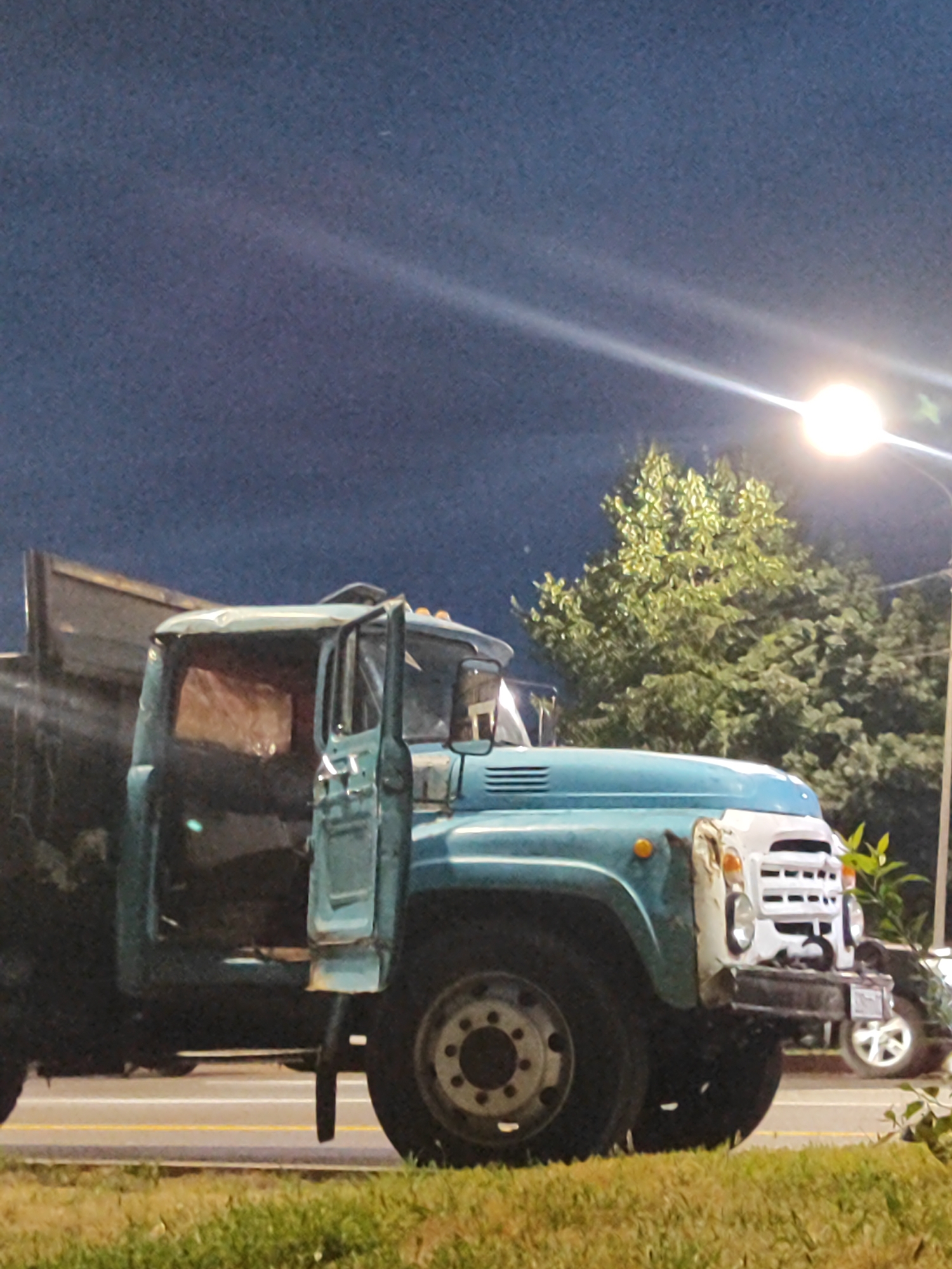 An old, teal and white truck is parked on the side of a road at night in Almaty, Kazakhstan. The truck has its driver's side door open and appears to be a dump truck. A bright street light illuminates the scene from the right, casting a strong glare and creating lens flare. Trees with lush green foliage are visible behind the truck, also lit by the street light. The sky is a deep blue, suggesting it is late evening or early morning. The road surface is visible with lane markings. A portion of another vehicle, possibly a car, is visible in the background on the right, its wheel and part of its body illuminated. The foreground consists of a grassy verge. There are no people visible in the image.