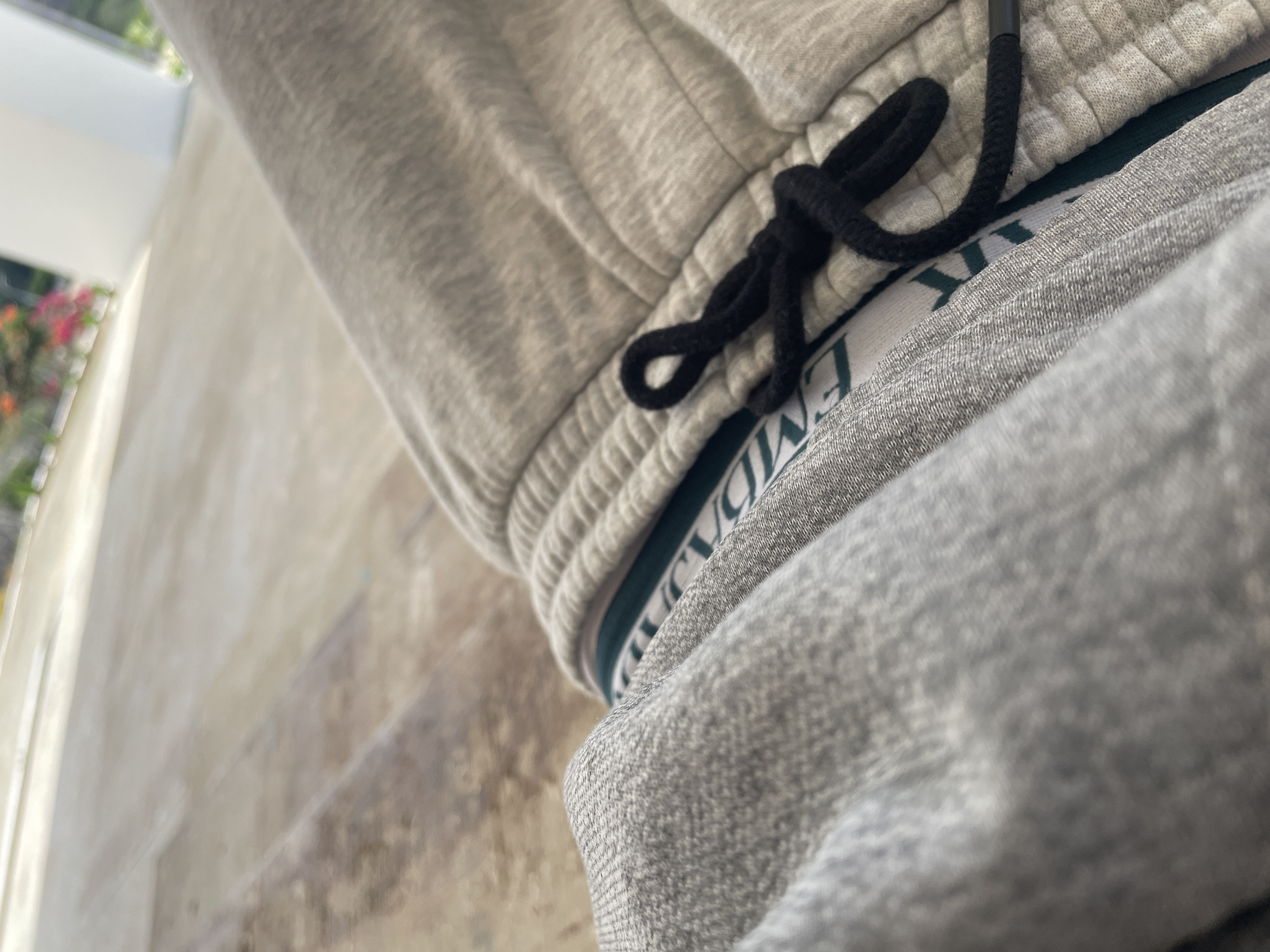 The image shows a close-up of a person's legs, wearing gray sweatpants over light-colored underwear with a dark green waistband featuring a repeating logo. A black drawstring is tied in a knot at the waist of the sweatpants. The background is blurred, showing a light-colored, textured surface that appears to be a patio or deck. In the far background, there is a glimpse of greenery and some pink flowers, suggesting an outdoor setting, possibly a balcony or garden area. The overall lighting indicates it is daytime. The El Valle del Espíritu Santo, Venezuela context is not directly depicted in the image.