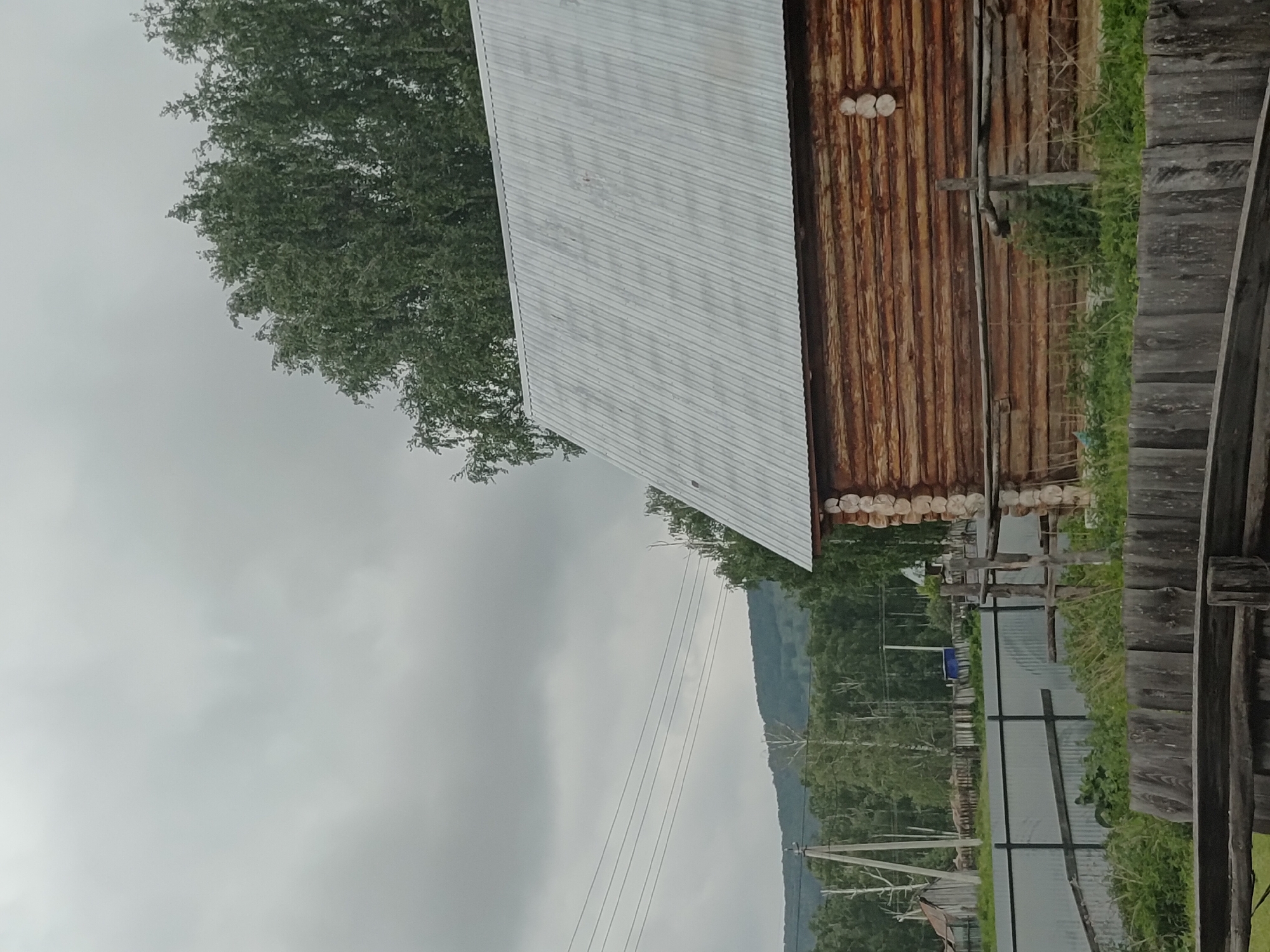 The image shows a rural scene on an overcast day, likely in Abzakovo, Russia. In the foreground, there is a weathered wooden fence. Behind it, to the right, is a portion of a rustic log cabin with a corrugated metal roof. The cabin's wooden walls are constructed from horizontally stacked logs. To the left of the cabin, a metal fence runs along the edge of a property, and beyond that, there are power lines strung between utility poles. Further in the distance, a dense forest covers rolling hills, with a treeline meeting the cloudy sky. The overall impression is one of a simple, rural dwelling amidst a natural landscape.