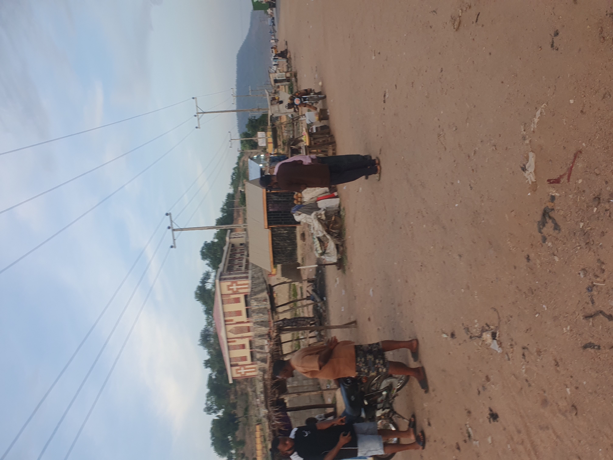 A wide, landscape-oriented photograph captures a slice of daily life along a dusty, unpaved road in Lokoja, Nigeria, under a bright, partly cloudy sky. The scene is set in what appears to be a semi-urban or rural area, characterized by simple structures and natural elements.
The foreground and midground are dominated by the reddish-brown dirt road, which shows scattered debris and small rocks. On the right side of the road, two young men are gathered around a motorcycle. One, dressed in a black t-shirt and light-colored shorts, is seated and appears focused on a device in his hands, likely a mobile phone. Further down the road, another man, wearing a light brown short-sleeved shirt and patterned shorts, stands casually next to a second motorcycle.
On the left side of the road, a man in a brown long-sleeved shirt and dark trousers stands with his back partly to the camera, near a series of rudimentary roadside stalls or shops. These structures, constructed from materials like wood and corrugated metal, suggest local commerce. Behind these stalls, a more developed two-story building with a red roof and distinct patterned walls is visible, surrounded by lush green trees.
In the distant background, a range of hills or mountains outlines the horizon, providing a geographical context for the location. Overhead, multiple power lines stretch horizontally across the sky, supported by wooden utility poles that line the left side of the road, indicating electrical infrastructure. The lighting suggests it is daytime, possibly late afternoon, with a soft, ambient glow. The individuals present appear engaged in casual, everyday activities. No discernible text is visible within the image.