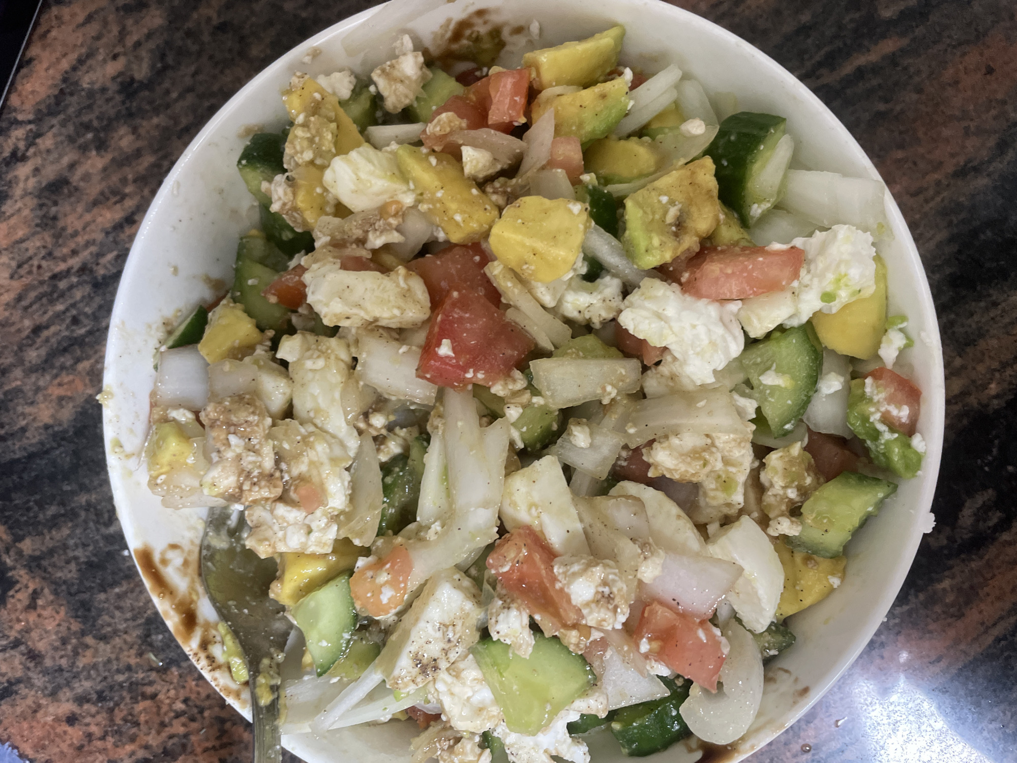 The image depicts a close-up view of a salad in a white bowl, likely photographed in a home setting. The salad contains cubed tomatoes, cucumbers, avocados, onions, and what appears to be feta cheese or a similar type of white cheese. A spoon rests within the bowl. The bowl sits on a dark brown countertop with a marbled pattern. The location of the image is likely in Petah Tikva, Israel.