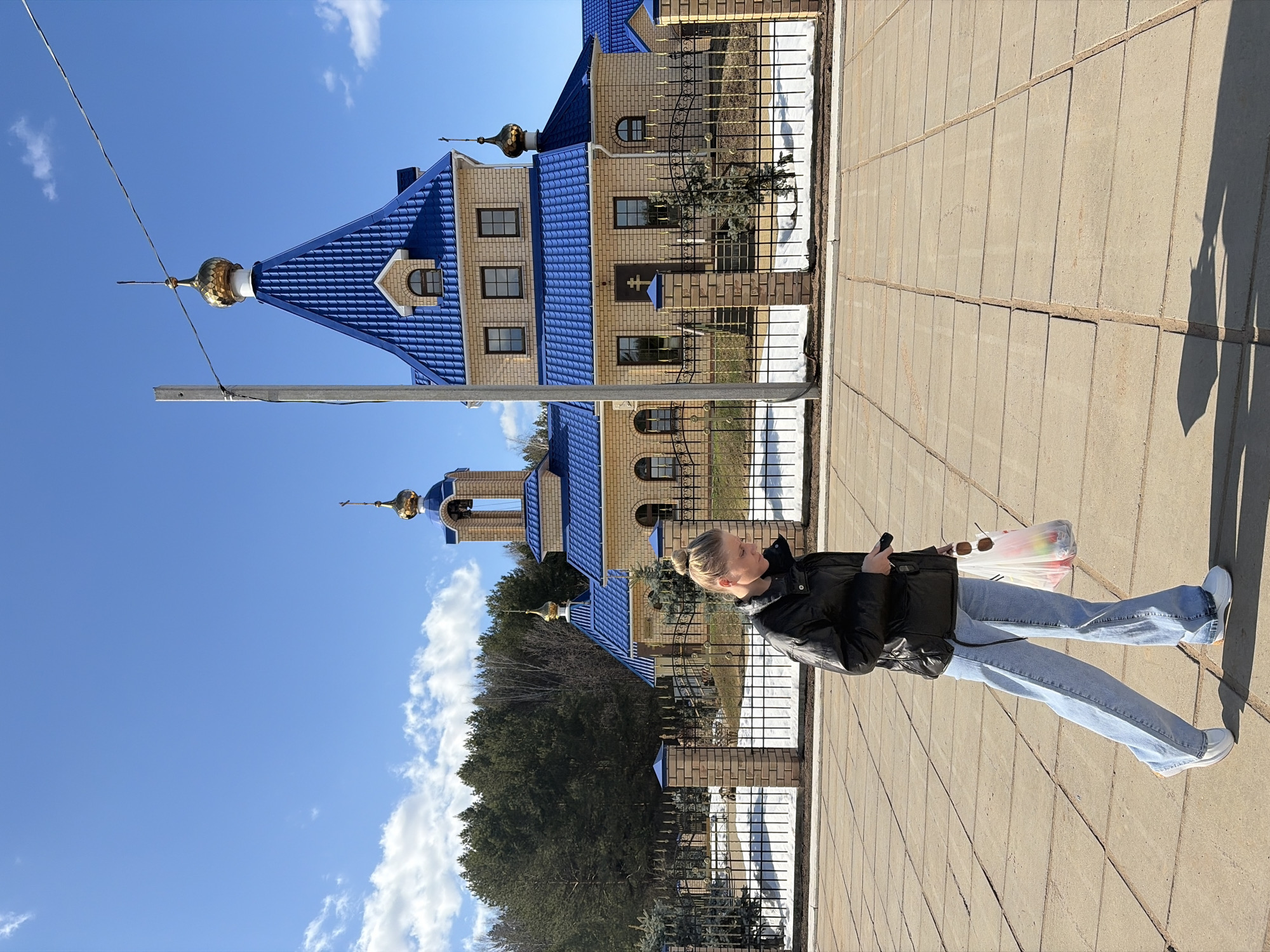 The image shows a young woman standing on a paved sidewalk in Chaikovsky, Russia. She is wearing a black jacket, blue jeans, and white shoes and is holding a shopping bag. In the background is the Church of the Kazan Icon of the Mother of God, a building with a blue roof, tan brick walls, and golden domes. The sky is blue with some clouds. The scene appears to be outdoors on a sunny day.