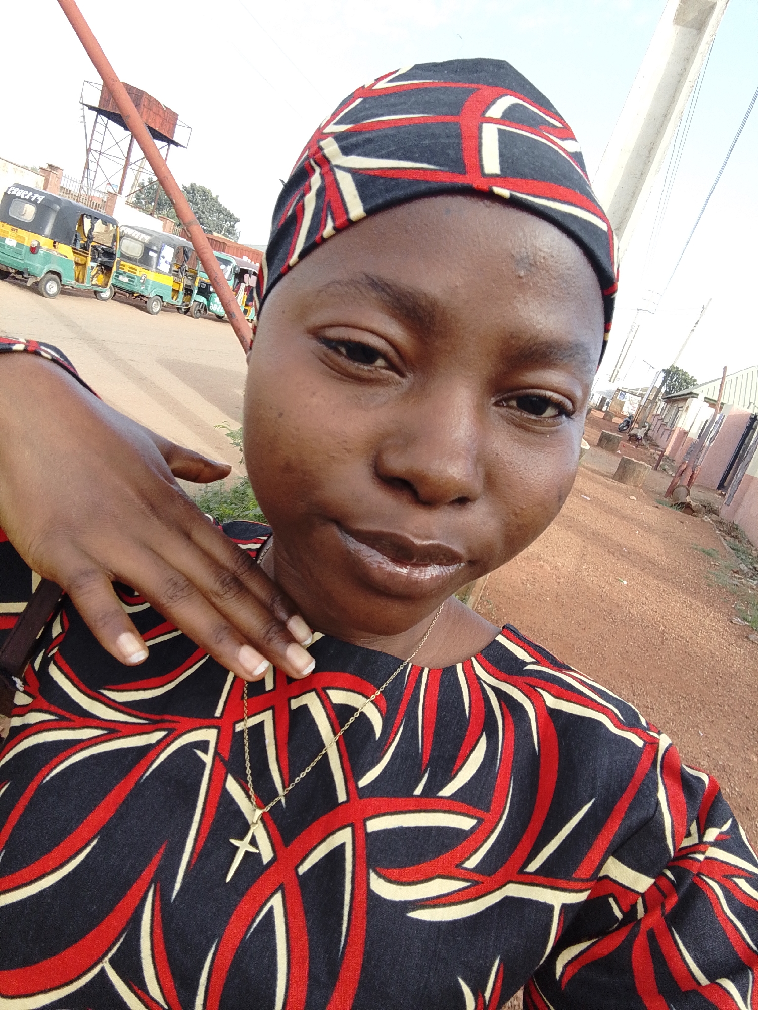 A medium, eye-level, outdoor shot of a dark-skinned young woman with a rounded face, black hair concealed under a dark hat with red and white geometric patterns, wearing a patterned black and red top, and a thin gold chain with a cross pendant. She gazes toward the camera with a slight, glossy-lipped smile, her hand resting against her neck. In the background, a row of green and yellow auto-rickshaws are parked on a street; behind these, a metal water tower is visible. The right side of the frame shows a dirt road adjacent to a building with a tan exterior and a white window frame. The location is speculated to be in the city of Jos, Nigeria, based on the presence of auto-rickshaws, also called "Keke Napep" in the background.