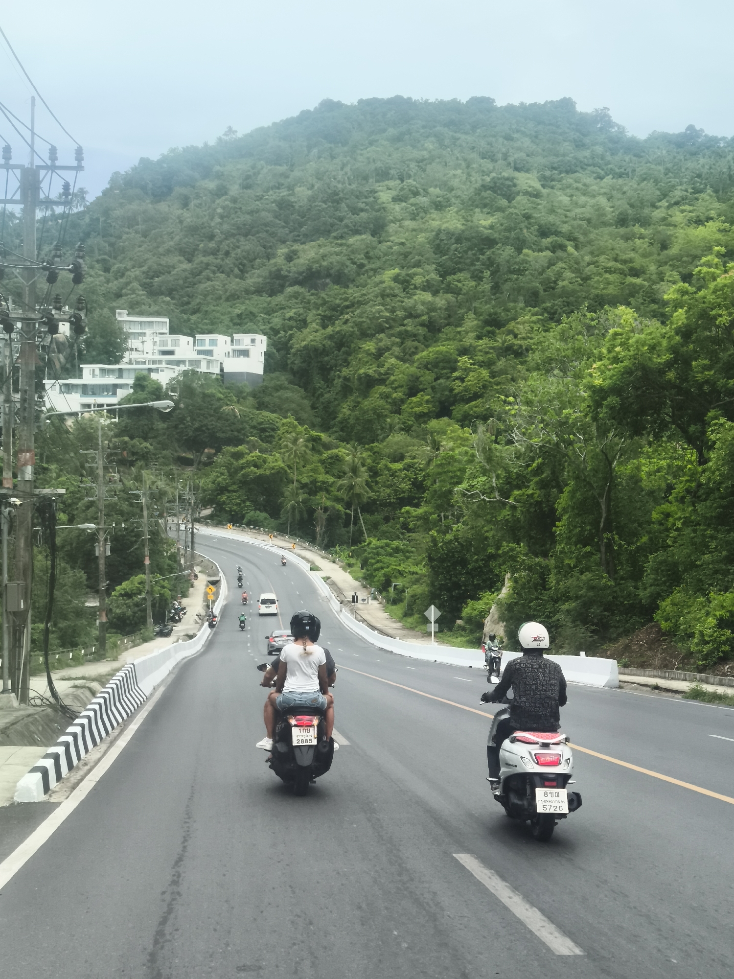The image depicts a scenic road winding through a lush, green, mountainous area in Baan Chaweng Noi, Thailand. In the foreground, two individuals are riding on a scooter, with the person in the back wearing a white t-shirt and denim shorts, and the person in front wearing a white helmet and a patterned black shirt. The scooter has a license plate with the number "2885". Further down the road, another person on a white scooter is visible, wearing a white helmet and a dark shirt. Several other vehicles, including cars and motorcycles, are present on the road. On the left side of the road, a series of utility poles with numerous wires are installed, and a modern white building complex with multiple levels is visible nestled into the hillside. The road is paved and features clear lane markings and a white and black striped barrier. The background is dominated by dense tropical vegetation covering the steep hillsides, under an overcast sky.