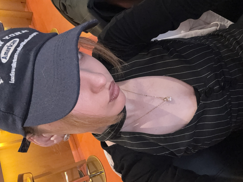 A close-up portrait depicts an individual seated in what appears to be an indoor establishment, possibly a bar or restaurant, located in Yerevan, Armenia. The exact venue cannot be determined from the visual information. The individual is wearing a dark-colored baseball cap, bearing the words 'NEW YORK' and 'American', pulled low over the face, obscuring the eyes. Light brown hair is visible around the face. The subject wears a black pinstriped top with a deep V-neck, a black choker, and a gold-colored necklace featuring a clear, heart-shaped pendant. A silver-toned stud earring is present on the visible left ear. The background features warm, orange-toned walls and a partial view of a gold-colored bar stool with a dark seat.