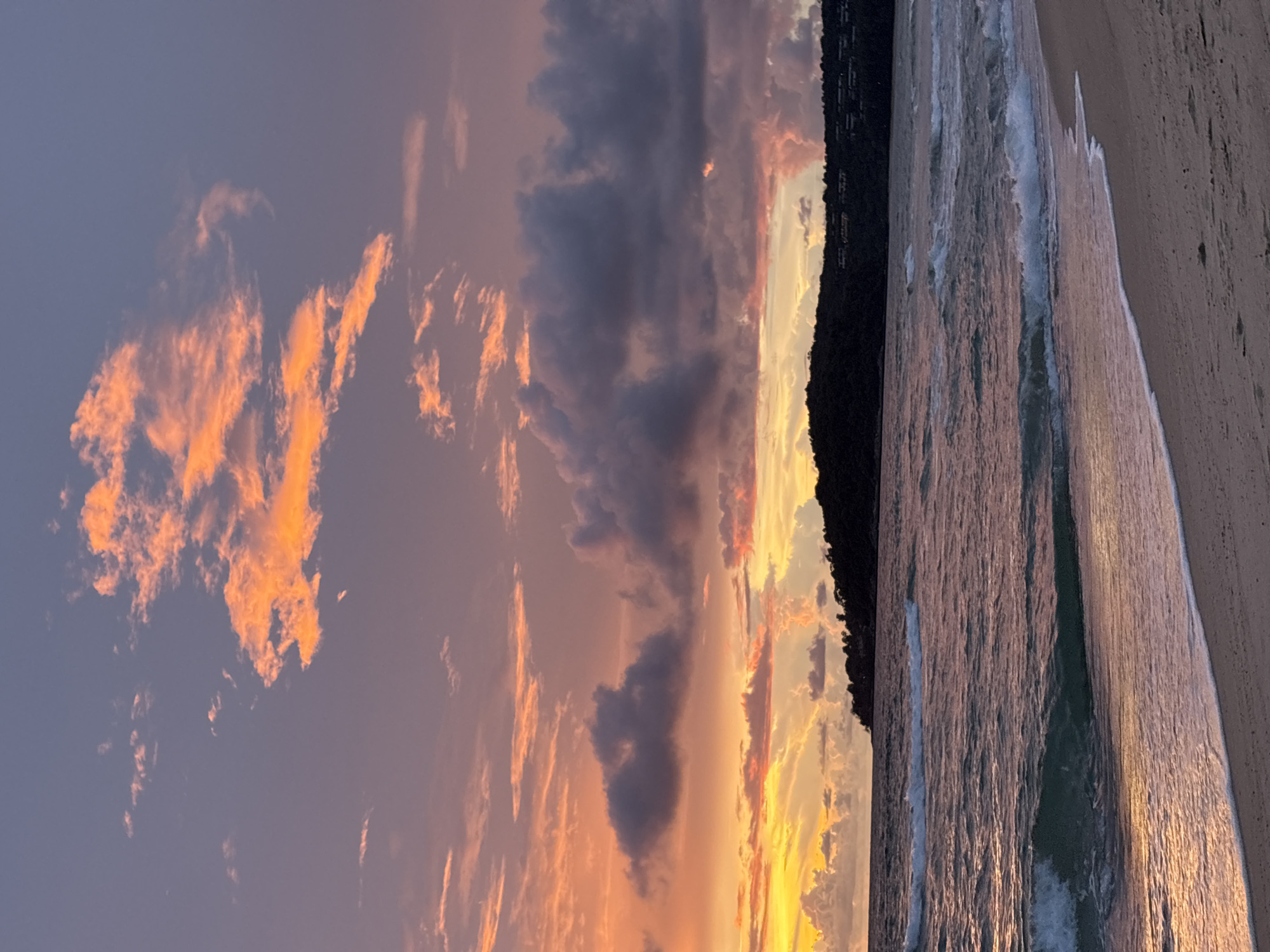 A sunset over a tropical beach in Choeng Thale, Thailand. The sky is filled with dramatic clouds painted in hues of orange, pink, and purple, illuminated by the setting sun. The ocean waves gently roll onto a sandy shore, reflecting the vibrant colors of the sky. A dark, tree-covered silhouette of a hillside borders the right side of the frame. No people or objects are visible in the scene. The image captures the serene beauty of a tropical evening.