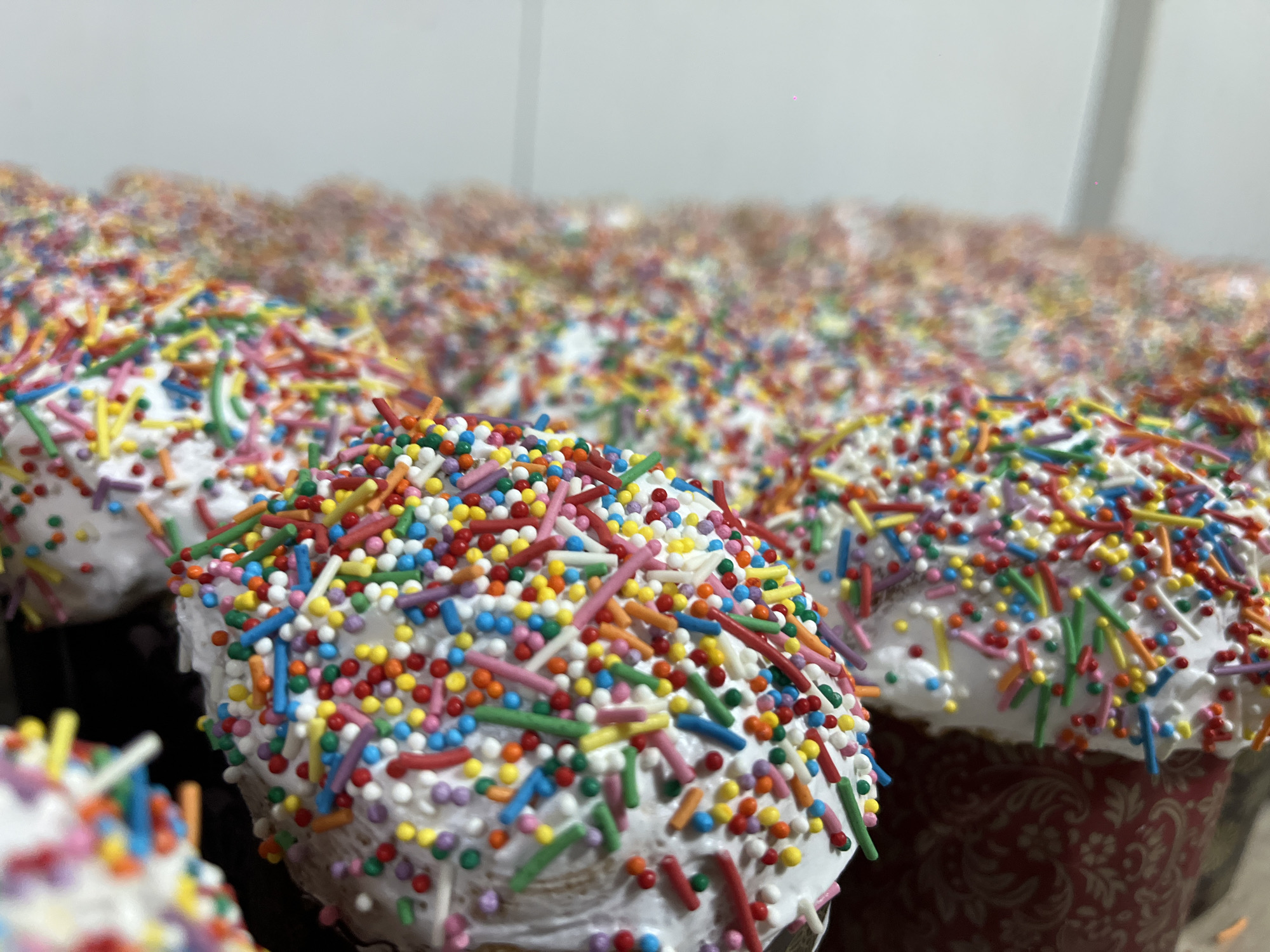 The image shows a large number of Easter cakes, known as Kulich, arranged in rows. The Kulich cakes are topped with white icing and decorated with colorful sprinkles. Some cakes are contained in decorative paper baking molds. The scene is indoors, possibly in Novopokrovskaya, Russia.