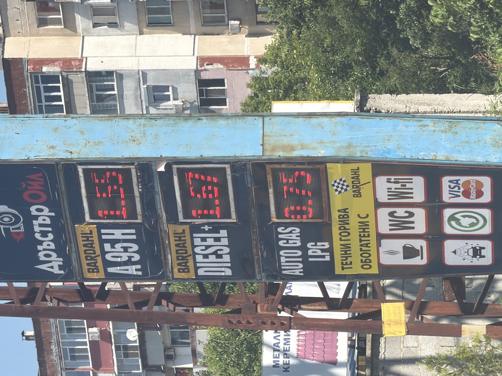 This media file, captured in Shumen, Bulgaria, depicts a gas station price sign (stele) and its surroundings under bright daylight. The main subject is a large, blue-framed sign displaying fuel prices and available services, mounted on a rusty metal support structure. In the background, a multi-story residential or commercial building with numerous windows is visible, along with lush green trees.
The price sign features the gas station's name in Cyrillic, "Дръстър ОЙЛ" (Drustar OIL). Fuel types and their prices are prominently displayed:
* A95H: 1.23 (likely Bulgarian Leva per liter)
* DIESEL+: 1.37 (likely Bulgarian Leva per liter)
* AUTO GAS LPG: 0.73 (likely Bulgarian Leva per liter)
The brand BARDAHL is visible next to the fuel types and on a yellow banner that reads "ТЕЧНИ ГОРИВА ОБОГАТЕНИ С" (Liquid fuels enriched with).
Below the fuel prices, various service icons and text indicate amenities offered:
* WC (restroom)
* Wi-fi
* Coffee cup icon (coffee/refreshments)
* Car wash icon
* VISA and MasterCard logos for payment
* A green recycling or environmental symbol.
In the lower part of the image, behind the sign's support structure, another sign is partially visible with the Cyrillic text "МЕТАЛИ КЕРЕМИ" (Metals Tiles). The overall scene suggests an urban setting with commercial activities.