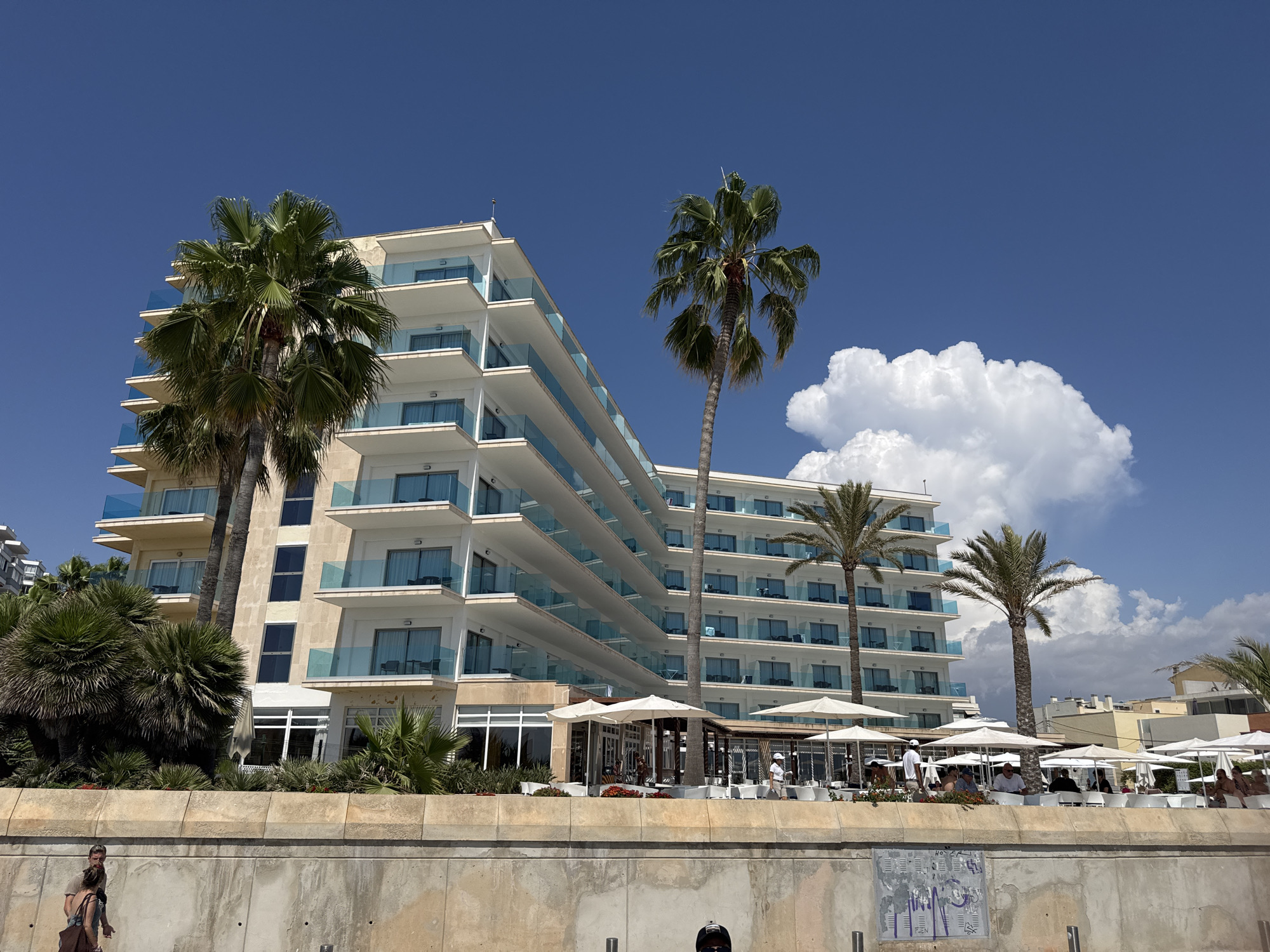 The image shows a modern, multi-story hotel with balconies and a beachside cafe area. Two prominent palm trees stand in front of the hotel. In the foreground, a concrete wall runs horizontally across the frame, with two people walking along the top of it. The sky is a clear blue with a few fluffy white clouds. The architecture of the hotel suggests it is located in a sunny, coastal region. Specifically, this hotel is the Melia Palma Bay, situated on the waterfront in Palma de Mallorca, Spain. The cafe area below the hotel is set up with numerous white umbrellas and tables, indicating it is a popular spot for guests and visitors. The people in the foreground appear to be casually dressed, enjoying a stroll along the promenade.