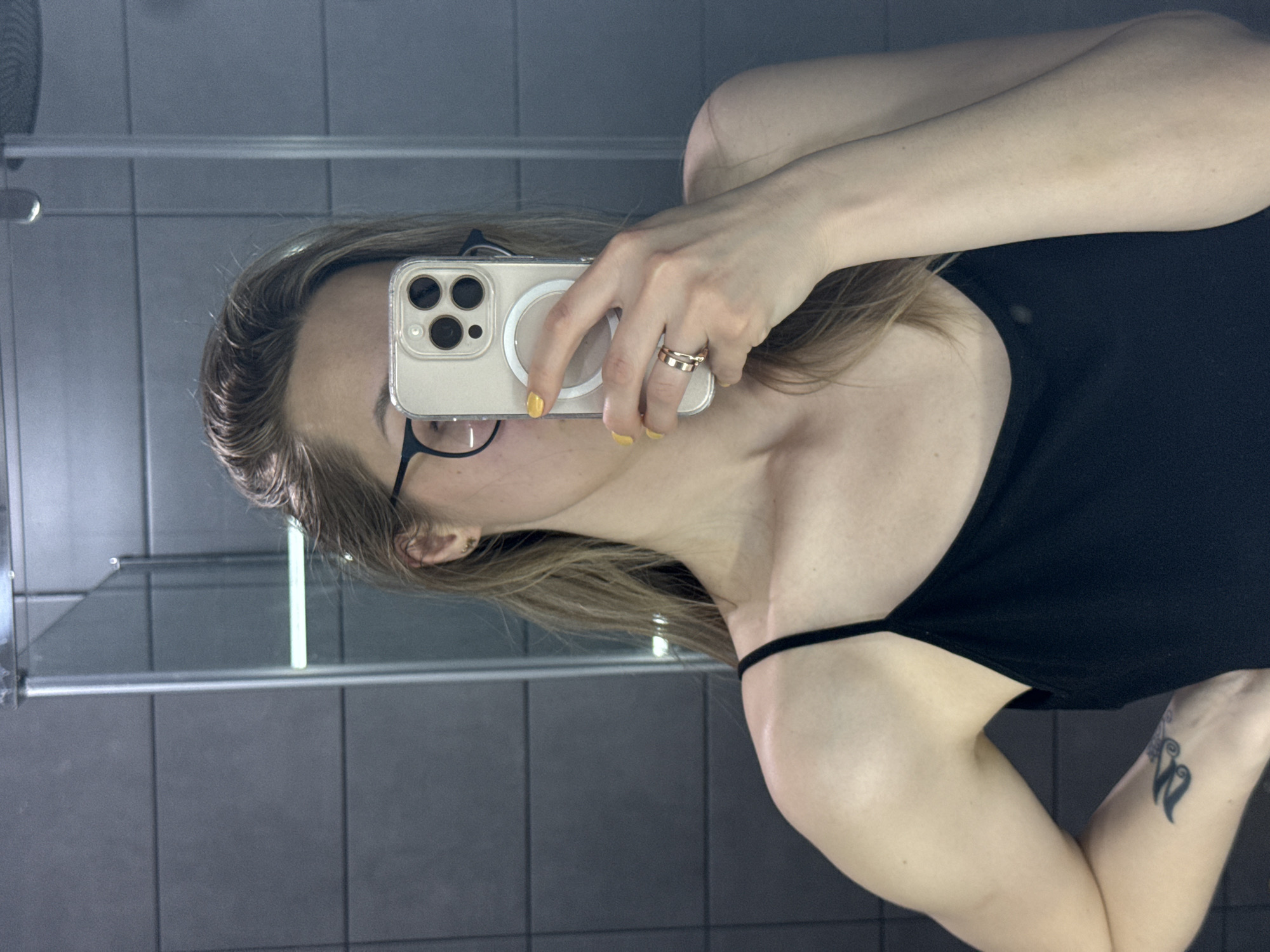 A person is taking a selfie in a bathroom mirror. The person is wearing glasses, a black tank top, and a ring on their finger. They are holding a smartphone with a beige case. The nails are painted yellow. The background shows a gray tiled wall, a shower rail, and a light fixture. There is a tattoo visible on the person's arm. The location is unknown and cannot be determined as being in the city of Chaikovsky, Russia.