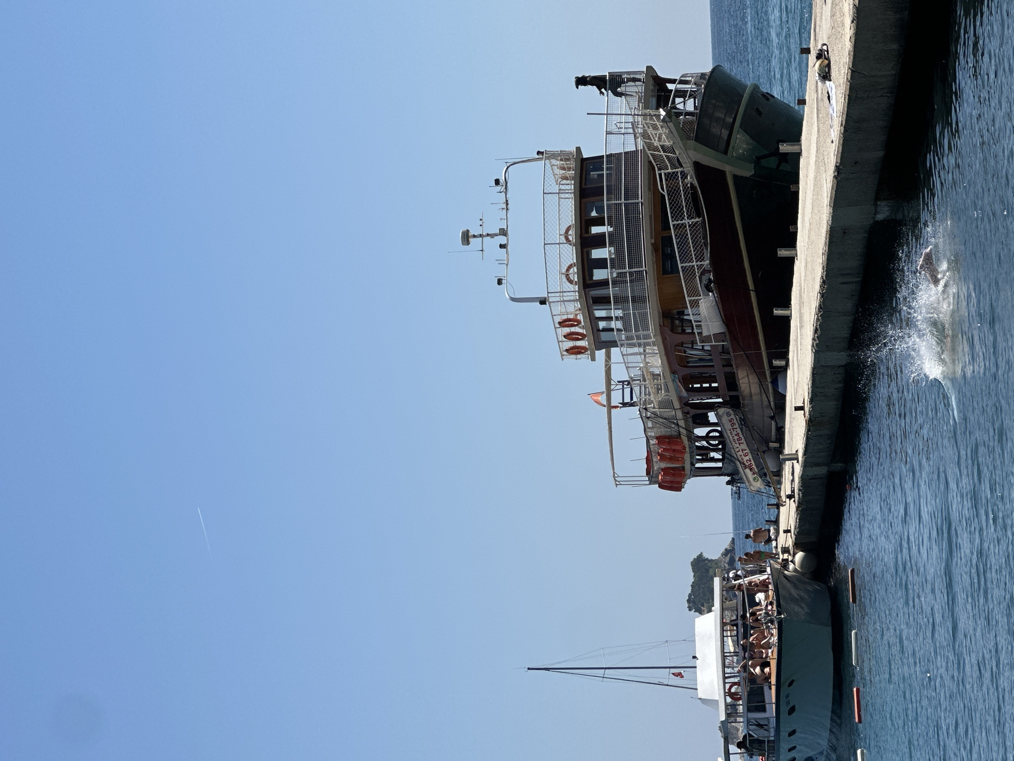 This image captures a sunny day at the Budva, Montenegro waterfront. In the foreground, the azure water is disturbed by a person jumping into it, creating a splash. Two boats are docked at a concrete pier. The larger boat, a multi-decked vessel with a reddish-brown hull and wooden accents, is positioned to the right. It features life preservers along its railing and a dark-clad figure standing on its upper deck. A sign with Cyrillic text and numbers is visible on its side. To the left, a lighter-colored, smaller boat is also docked, carrying several passengers lounging on its deck. A sailboat with a tall mast stands behind this boat, with a small, rocky island visible in the distance. The sky above is a clear, pale blue with a faint contrail from an airplane.