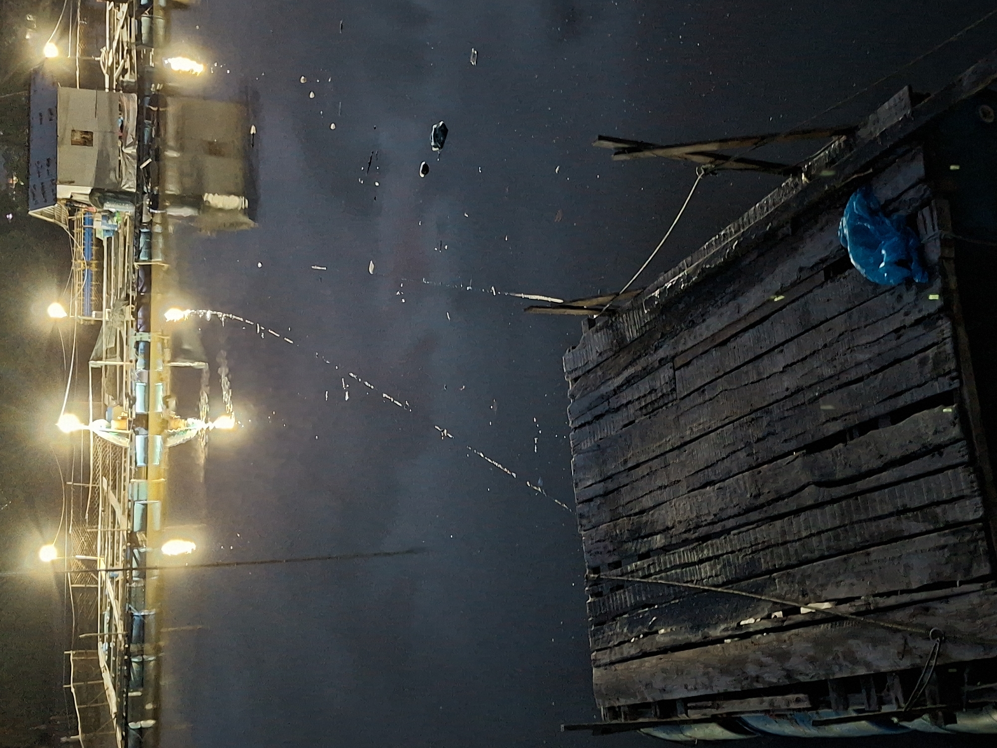 A nighttime scene depicting a wooden structure extending over the water. The structure is built of planks and beams, with a blue bag visible on one side. To the left, a tall structure with lights stands along the edge of the water; its reflection is visible. The location appears to be the Krueng Cunda River in Lhokseumawe, Indonesia, based on the reflection in the water and the architectural styles. The water is dark and shows some scattered reflections and floating debris.