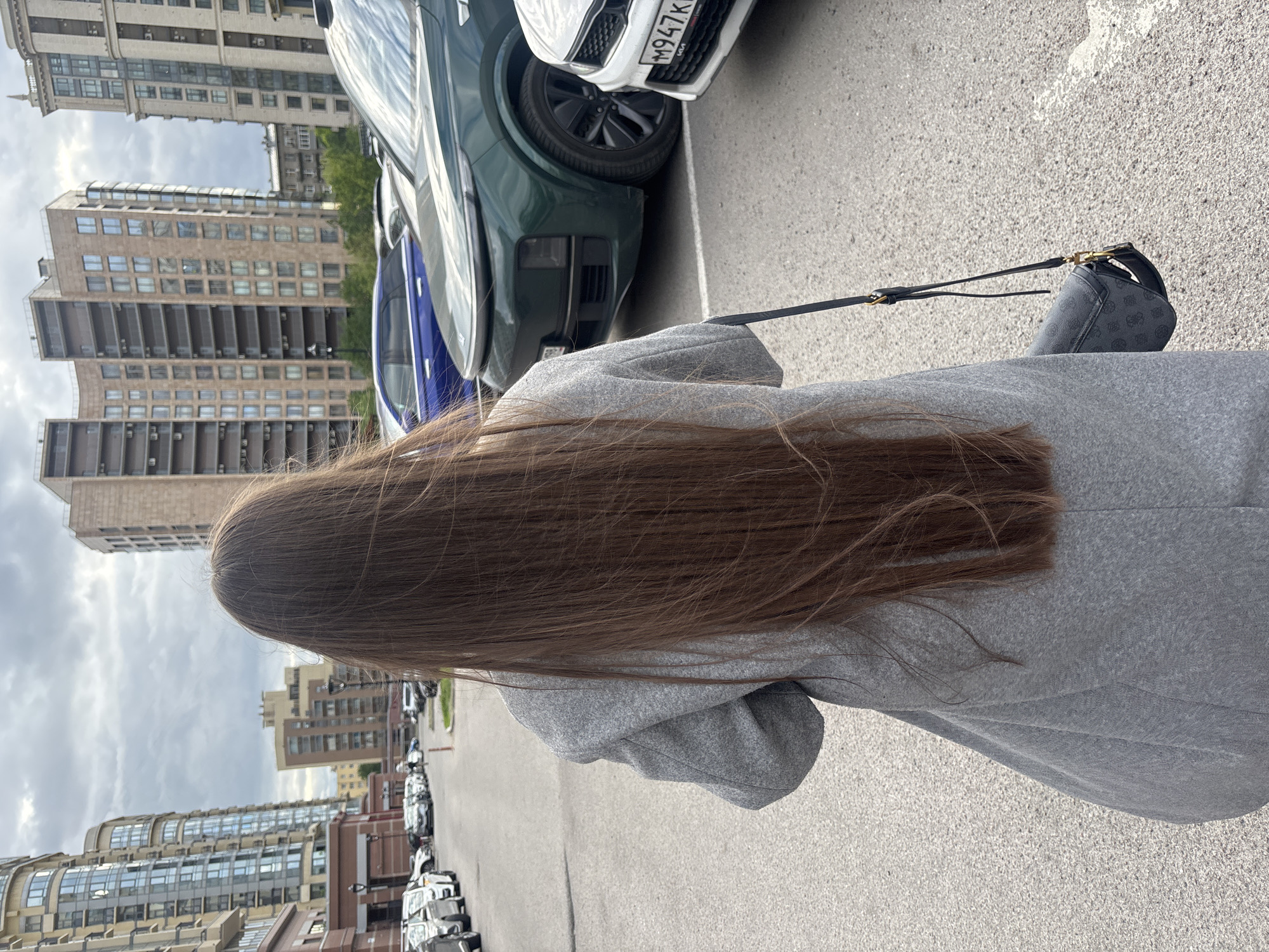 A photo taken at street level depicts the back of a person with long brown hair, wearing a gray coat, and holding a small black purse. Several cars are parked nearby, and buildings are visible in the background. The photo appears to have been taken in a city environment.