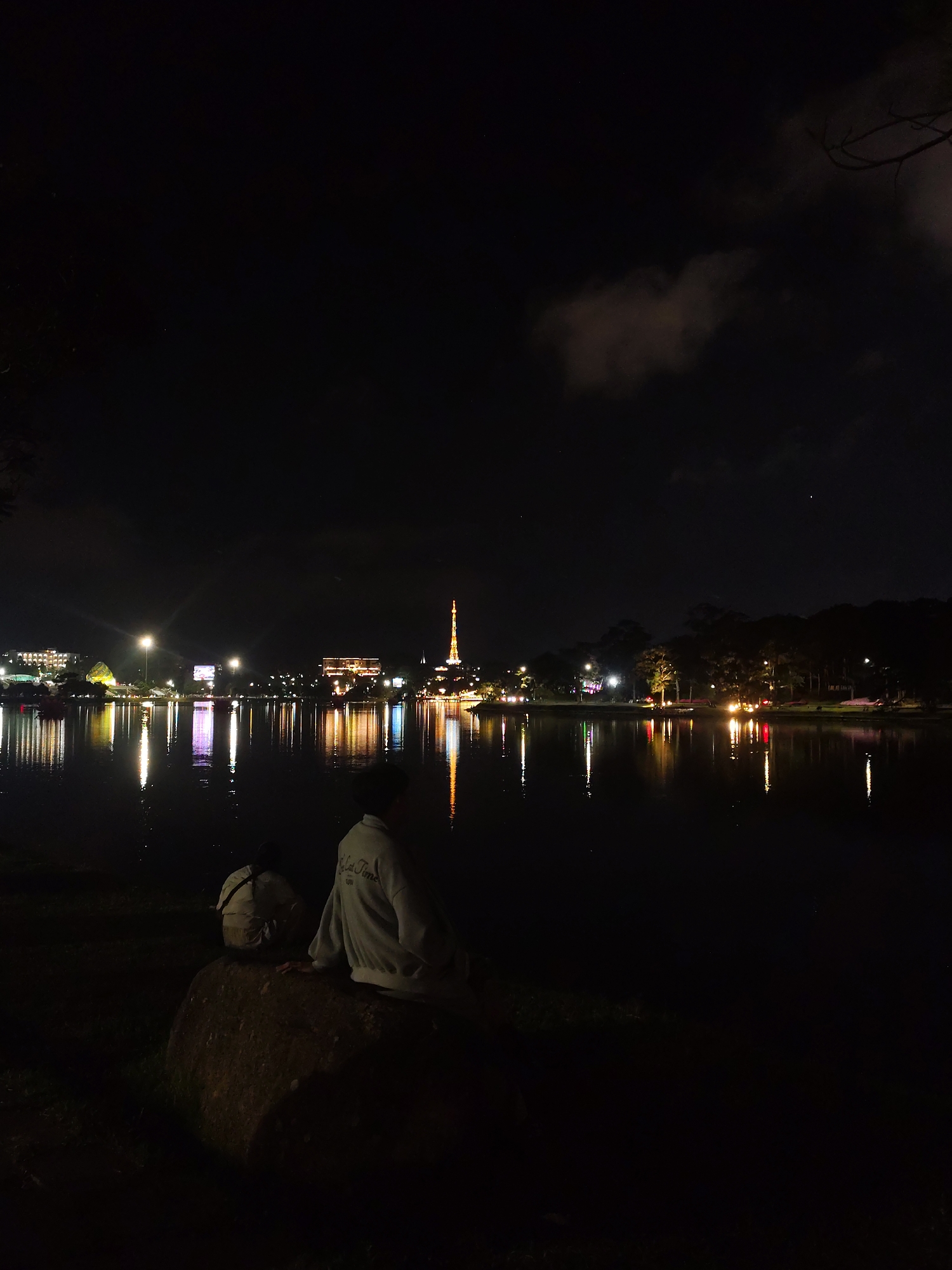 The image depicts a night scene at Xuan Huong Lake in Da Lat, Vietnam. Two people are sitting on a large rock at the edge of the lake. One is crouched lower, and the other is leaning slightly forward with one hand on the rock. Reflected lights from buildings and a prominent tower are visible across the water. The background includes a city skyline, trees, and clouds in the sky. The scene is dimly lit with light sources from the city and faint starlight.