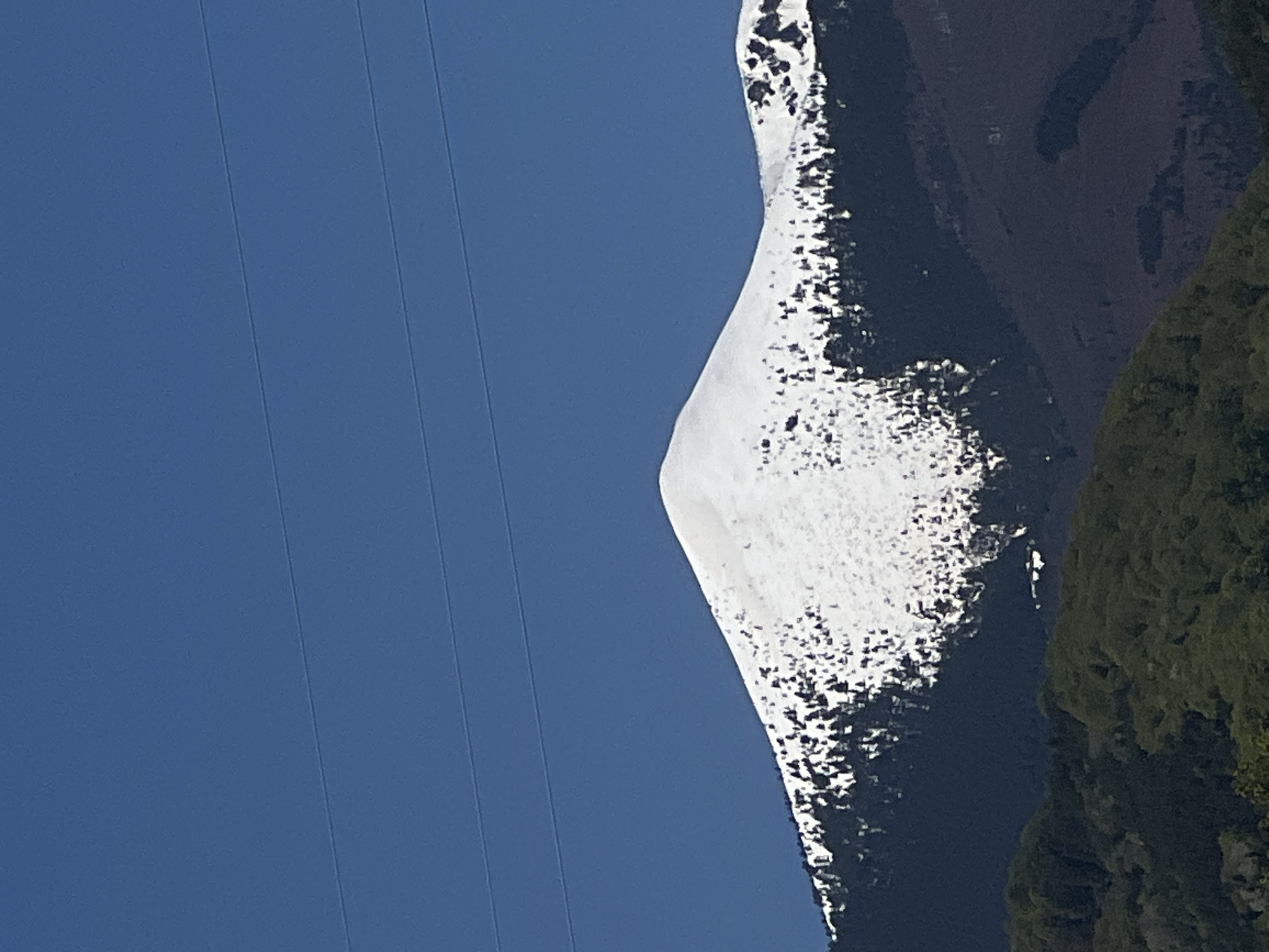 The image shows a portion of a mountain landscape taken from a low angle, the mountain is covered in snow with some exposed dark rock and vegetation. In the sky, there are visible power lines and a clear blue sky. This scene is located in or near Kornitsa, Bulgaria.