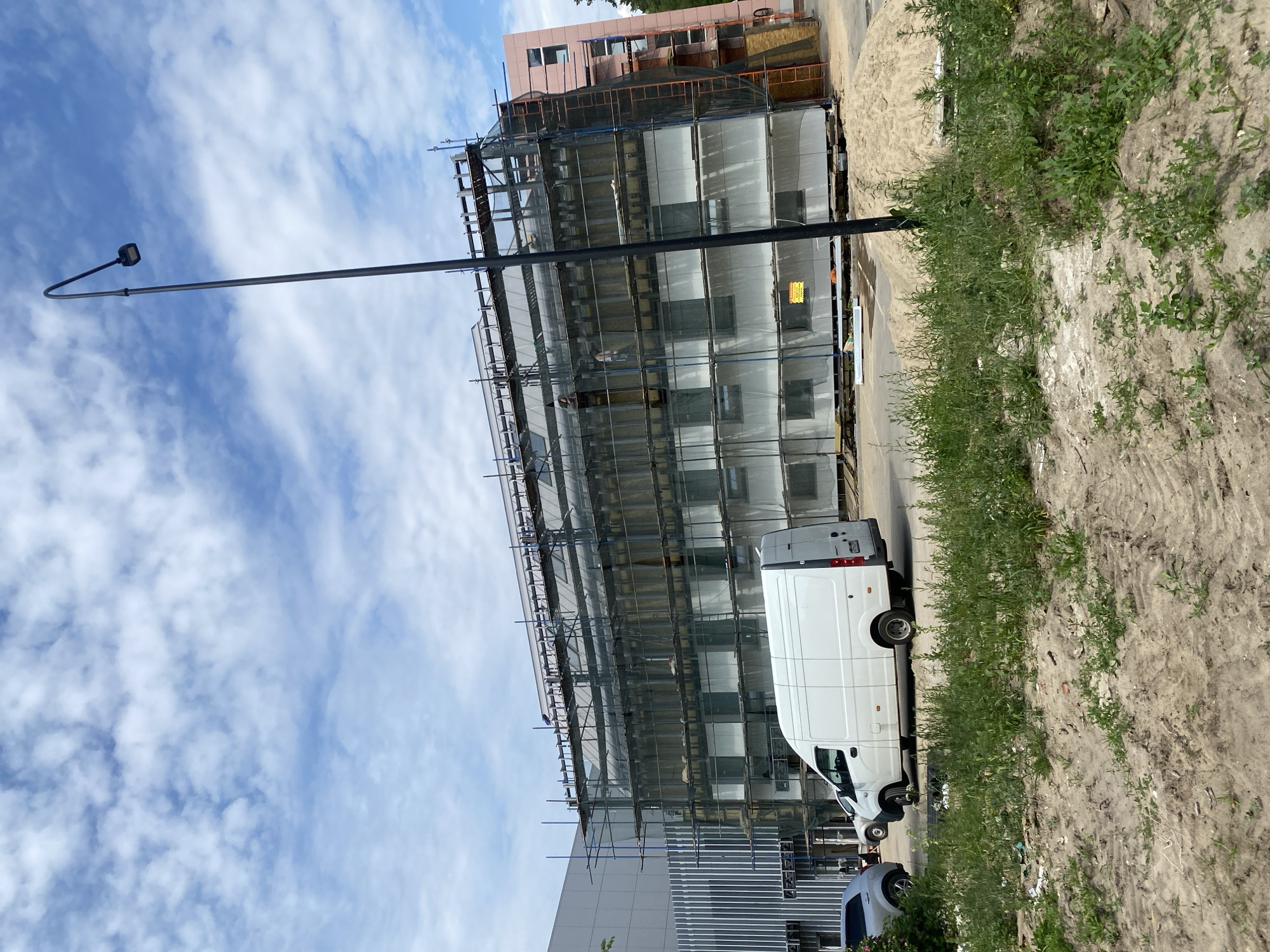 An exterior, eye-level, full shot shows a building under construction in an urban area under a partly cloudy sky. The building is clad in white panels and covered in scaffolding, with a safety net over the scaffolding. To the left of the building is a large, modern gray building with vertical metal siding. A white van is parked in front of the construction site, and to the left of the van, a silver car is partially visible. The foreground is a patch of dirt and weeds. A tall black lamppost stands to the right of the building, and a pile of sand is visible next to the construction site.