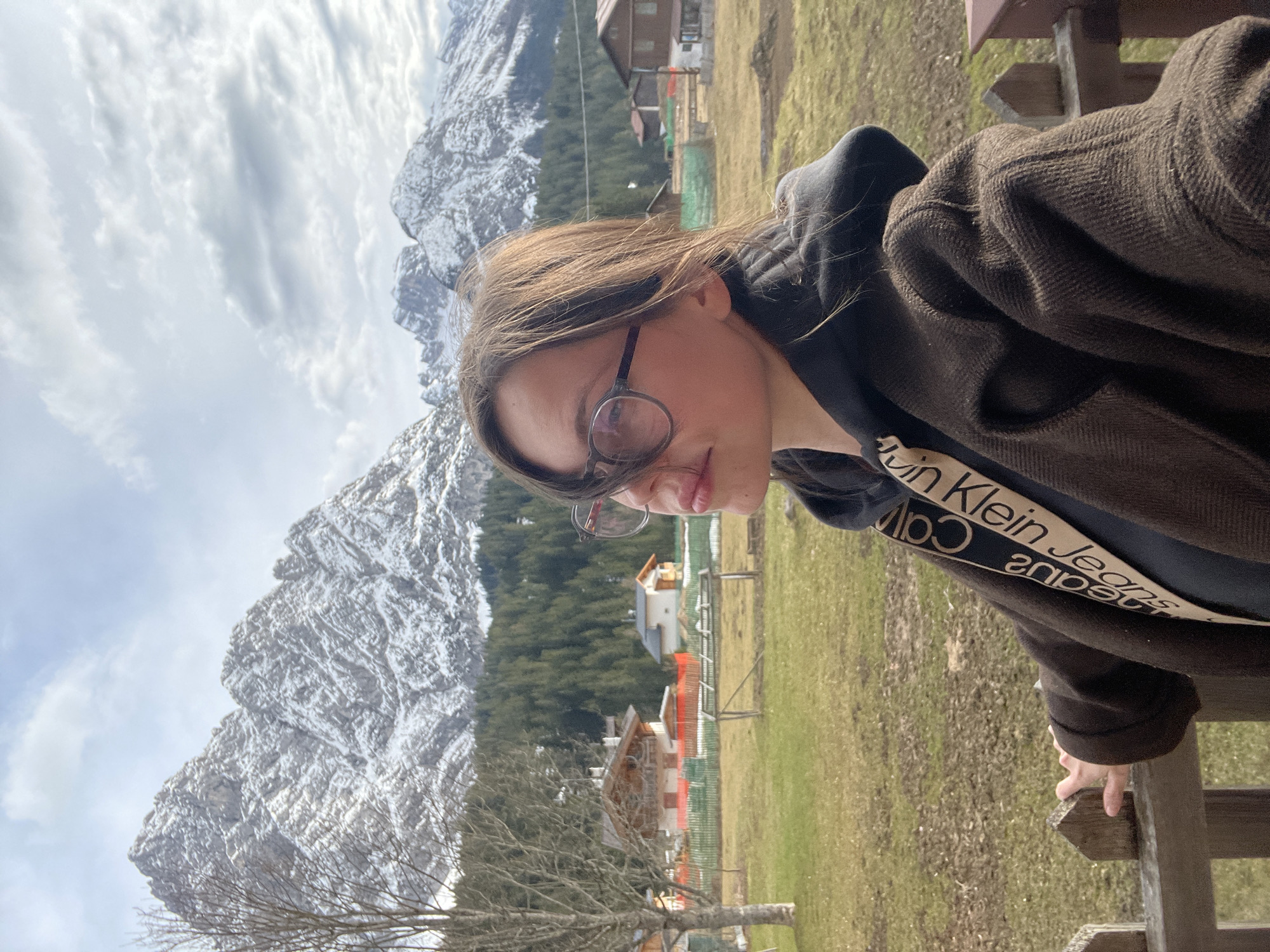 A person with glasses stands near a wooden fence. They are wearing a black hoodie with a Calvin Klein Jeans band and a brown jacket. In the background, there is a mountain range partially covered in snow, a field, and buildings. This scene appears to be in Sappada / Plodn / Sapade, Italy.