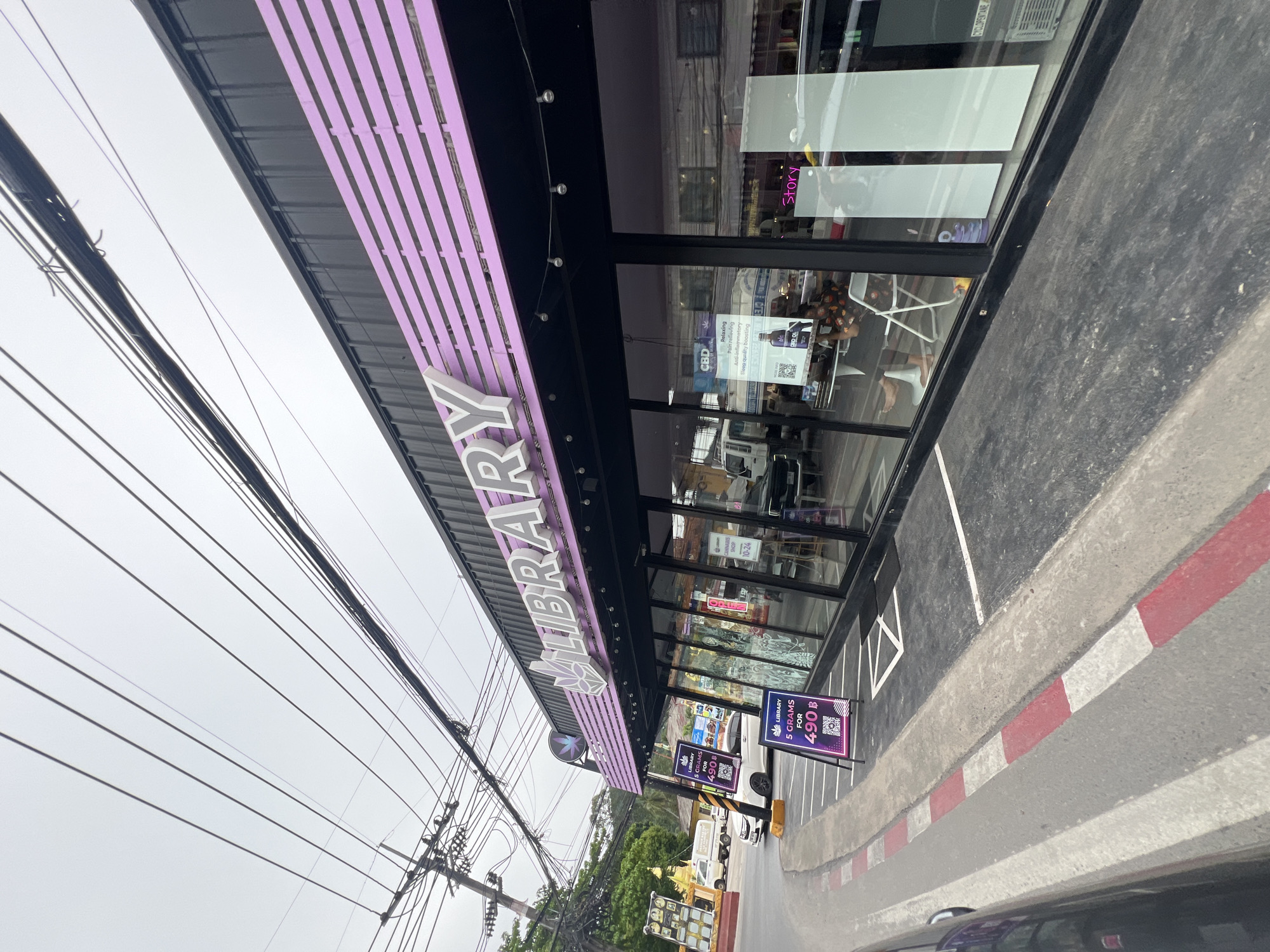 The image shows the exterior of a cannabis shop named "Library" in Baan Hua Thanon, Thailand. The shop has a black facade with a prominent "LIBRARY" sign in large, white, 3D letters, outlined in purple. A purple decorative element with a leaf design is positioned to the left of the sign. The shop features a large glass window front, through which the interior is visible. Inside, there are tables, chairs, and displays. Two purple A-frame signs are placed on the sidewalk in front of the shop, advertising "LIBRARY 5 GRAMS FOR 490 B" along with QR codes.
The street in front of the shop has a road with a curb marked with red and white stripes. Power lines are visible overhead, crossing the sky. In the background, there are trees, other buildings, and vehicles, including a white truck and a white car parked near the shop. The sky is overcast. A person is seated inside the shop, visible through the glass.