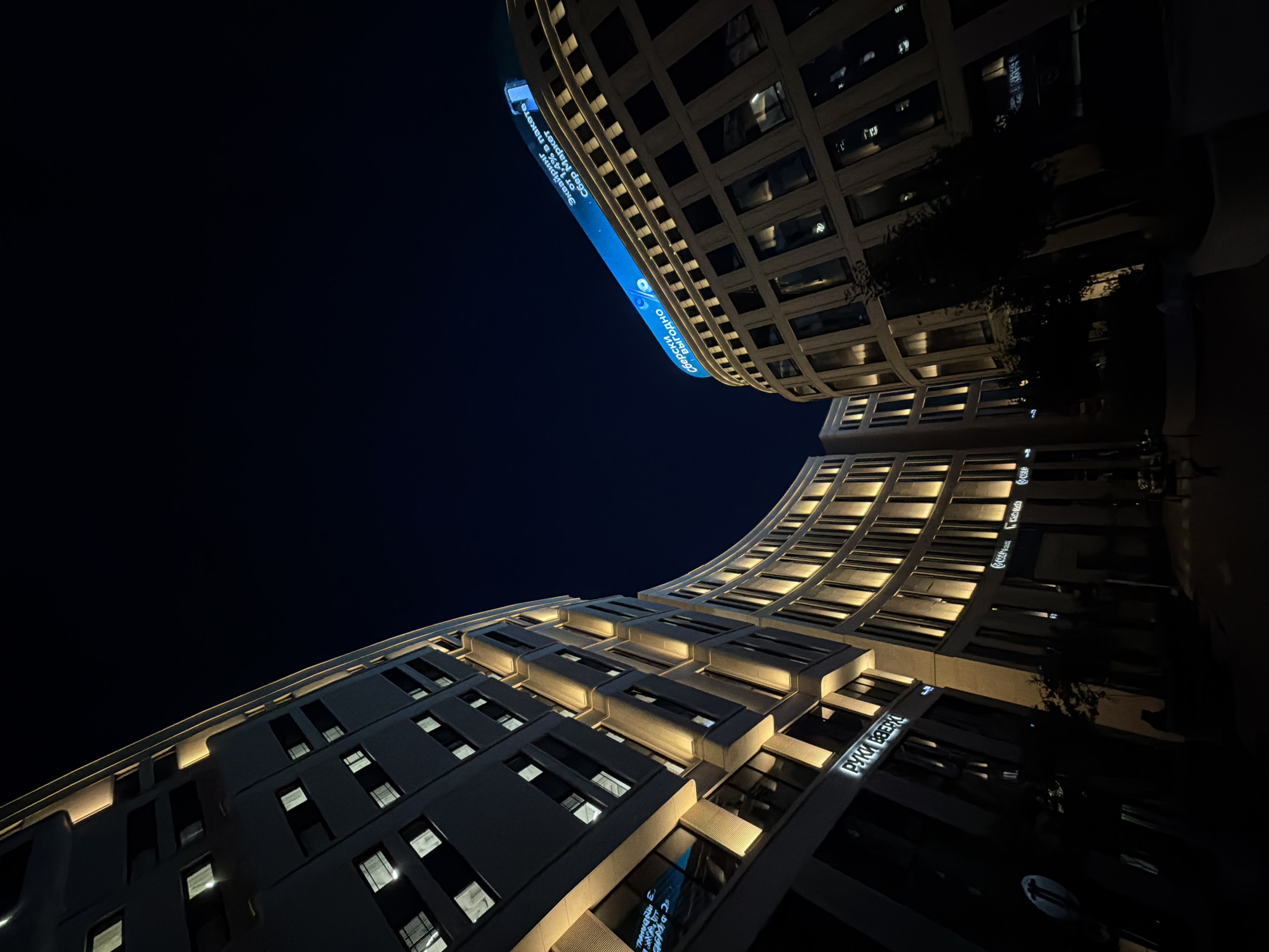 The image captures a night view of a modern architectural complex in Minsk, Belarus, featuring two large, curved buildings illuminated by warm exterior lighting. The left building, with its light-colored facade, displays numerous rectangular windows, some of which are lit from within, suggesting interior activity. Yellowish lights accentuate the vertical lines of this structure. A sign visible on the lower section of this building reads "РУКИ ВВЕРХ".
The building on the right is larger and features a more uniform facade with a grid of dark windows. The top of this building is adorned with a prominent, bright blue illuminated sign that displays Cyrillic text, reading "Сберки выгодно" and "Эквайринг от 1.4% в пакете Сбер Маркет". This suggests a commercial or financial district. The overall scene is characterized by the contrast between the dark night sky and the illuminated architecture. There are no discernible people or specific actions depicted in the image. The perspective is from a low angle, looking upwards towards the sky, emphasizing the scale and grandeur of the buildings.