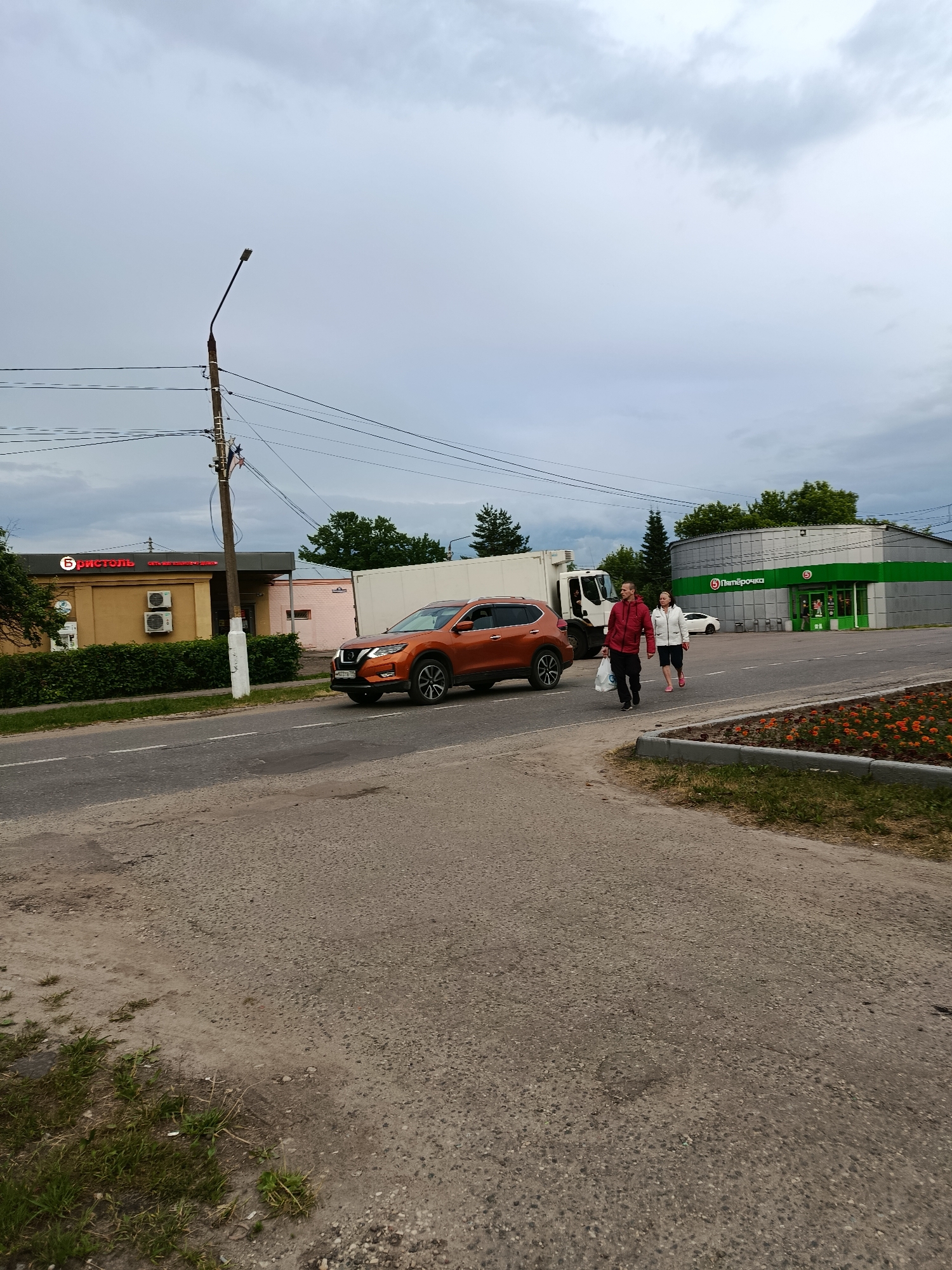 In the city of Veraya, Russia, on a cloudy day, an orange Nissan Qashqai is parked on the right side of the road, partially on the shoulder. Further down the road, a white truck is visible. Two individuals, a man in a red jacket and a woman in a white jacket, are walking across the road from right to left, carrying a white plastic bag. To the left of the road, a yellow building with red lettering and air conditioning units is visible, along with a sidewalk and a neatly trimmed hedge. To the right of the road, a green and silver building with the "Пятёрочка" logo is present, suggesting it is a grocery store. The foreground features a road with a visible pothole and patches of grass on the side.