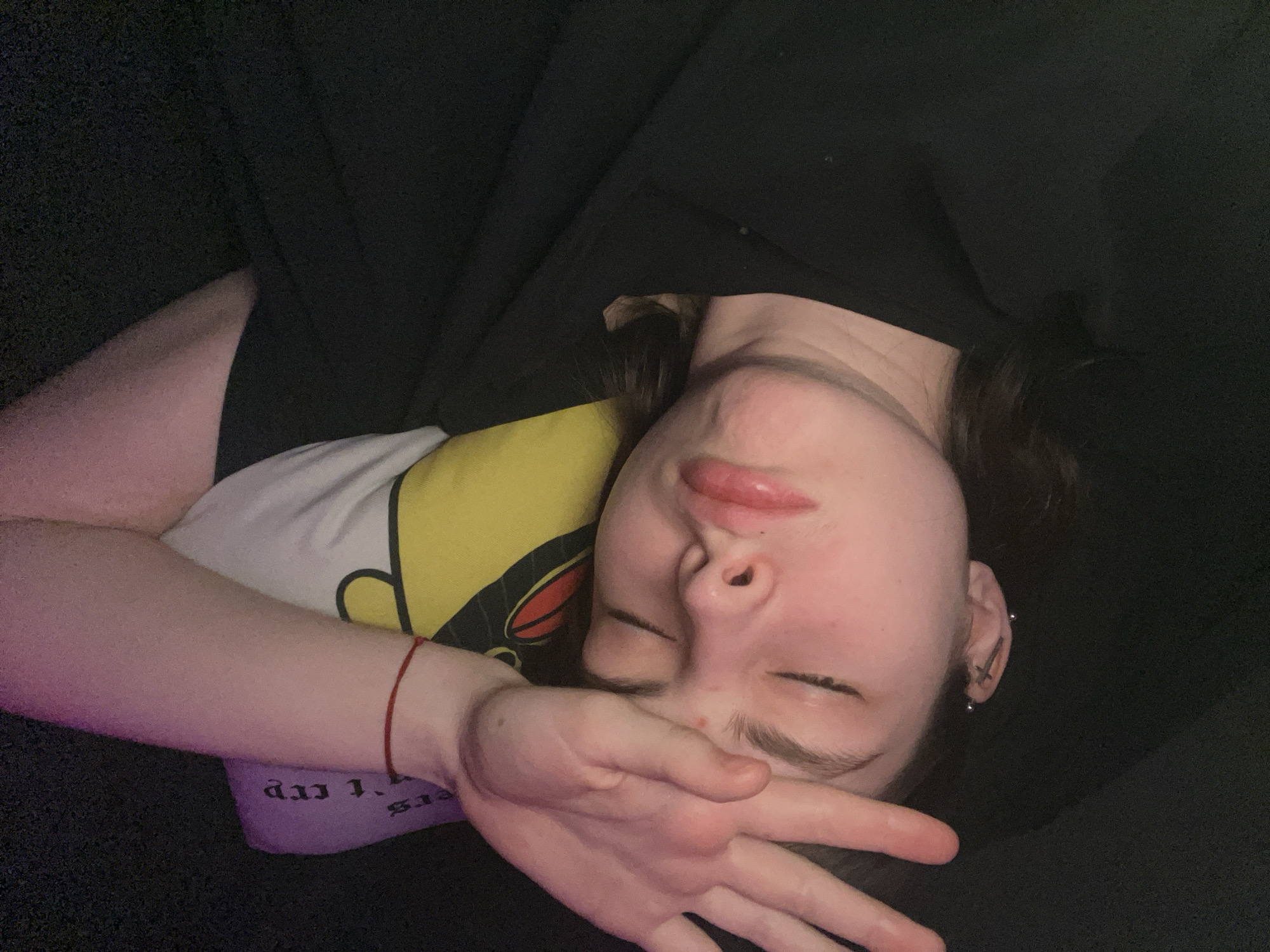 A person is lying on their back on a dark surface, possibly a couch or bed. Their face is partially obscured by their right hand, which is held up in front of their face. They have closed eyes and a slightly pursed mouth. The person is wearing a t-shirt with a cartoon graphic and a black top. They have multiple piercings in their left ear. A red string bracelet is on their left wrist. No information suggests this photo was taken in Saint Petersburg, Russia.