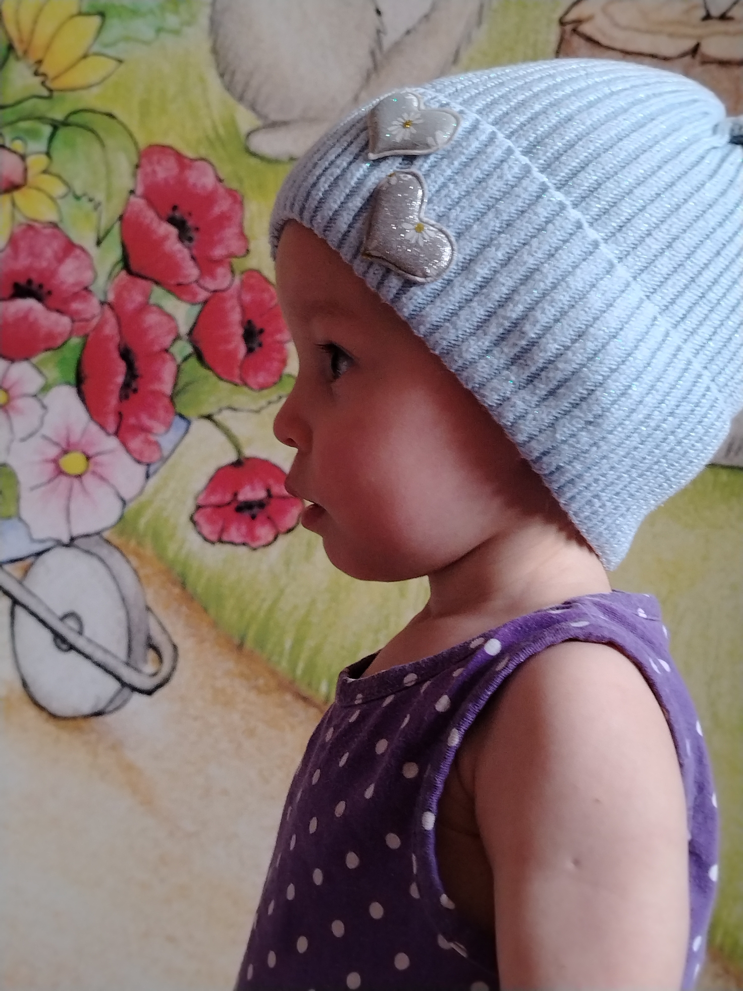 A young child, likely a toddler, is captured in a profile view looking towards the left side of the frame in an indoor setting in Chaikovsky, Russia. The child wears a light blue ribbed beanie adorned with two silver heart-shaped embellishments, each featuring a small white daisy design. Their lips are slightly parted, and their gaze is directed with an air of curiosity or focus. The child is also wearing a purple sleeveless dress with white polka dots.
The background is a vibrant and colorful mural or illustration. On the left, there is a cluster of prominent red poppies with dark centers, alongside pink and yellow flowers. Below these, part of a white wheelbarrow or cart with a visible wheel and handle can be seen. The ground is depicted with green grass, and in the upper right background, a partial view of a white, cartoon-like animal, possibly a rabbit, is visible. The bright lighting suggests it is daytime. There is no visible text in the image.