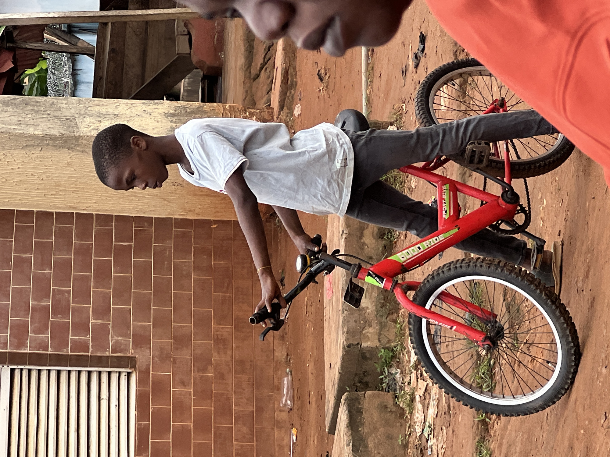 An outdoor, medium shot captures a young Black boy with short, dark hair, wearing a light blue t-shirt and dark gray pants, standing next to a red bicycle. He is holding the bicycle's handlebars with both hands, his right hand grasping the grip and brake lever. The bicycle has a black seat, black pedals, and black tires with white rims. The frame of the bicycle has "SPORT BIKE" written on it in green lettering. The boy is standing on a dirt ground in what appears to be a neighborhood in Nkpor, Nigeria. Behind him is a brown brick building with a window that has horizontal white louvers. To the right of the boy, another Black person, out of focus, is partially visible in the foreground, wearing an orange shirt. The background includes more buildings and some vegetation. The lighting suggests it is daytime.