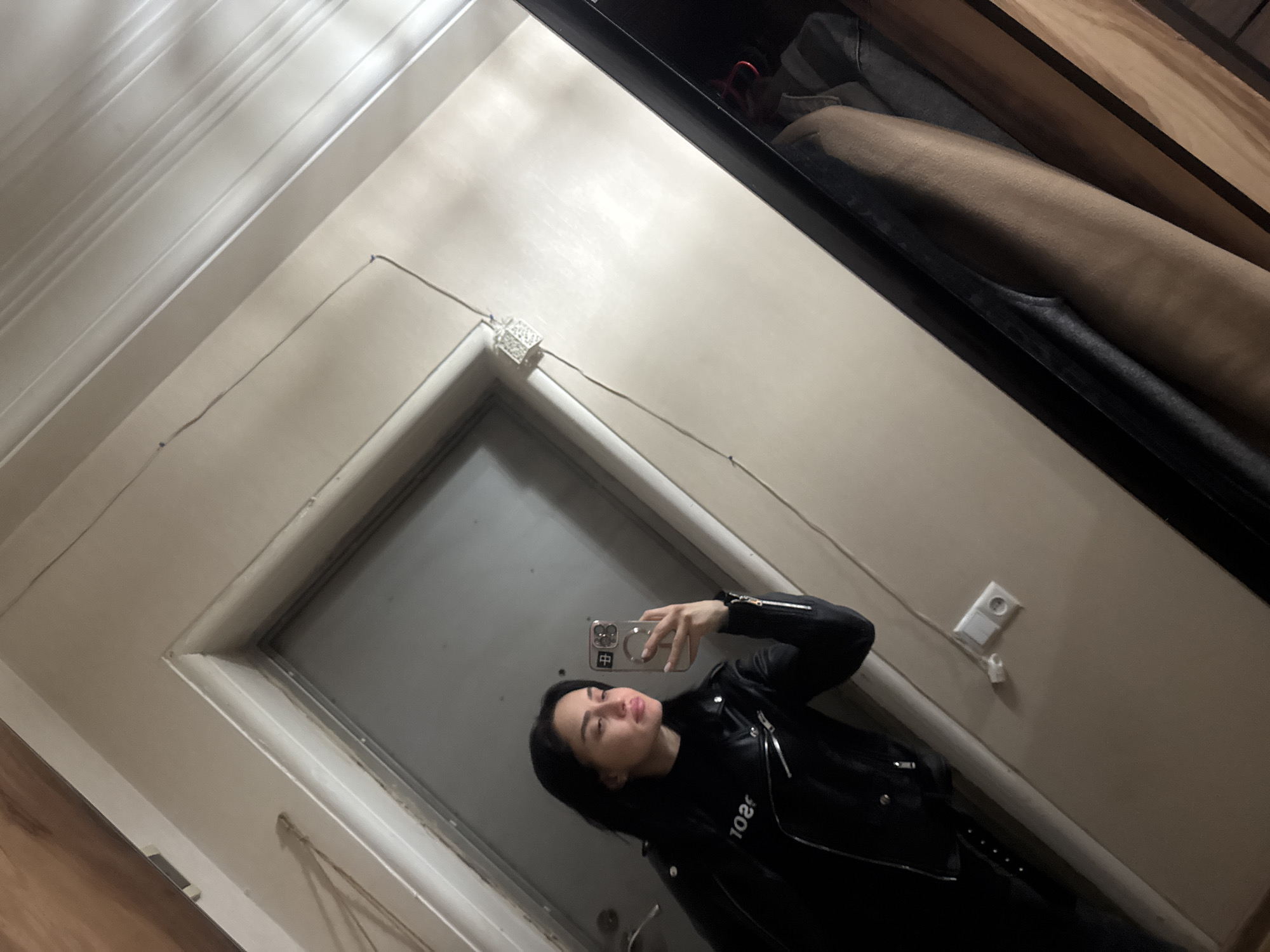 A young woman is taking a selfie in a mirror. She is wearing a black leather jacket. The location appears to be inside a building in Tbilisi, Georgia, as indicated by the Cyrillic characters on the phone case. The room has white walls and a door. There is an electrical outlet and a junction box on the wall. A closet is visible in the upper right corner of the mirror.