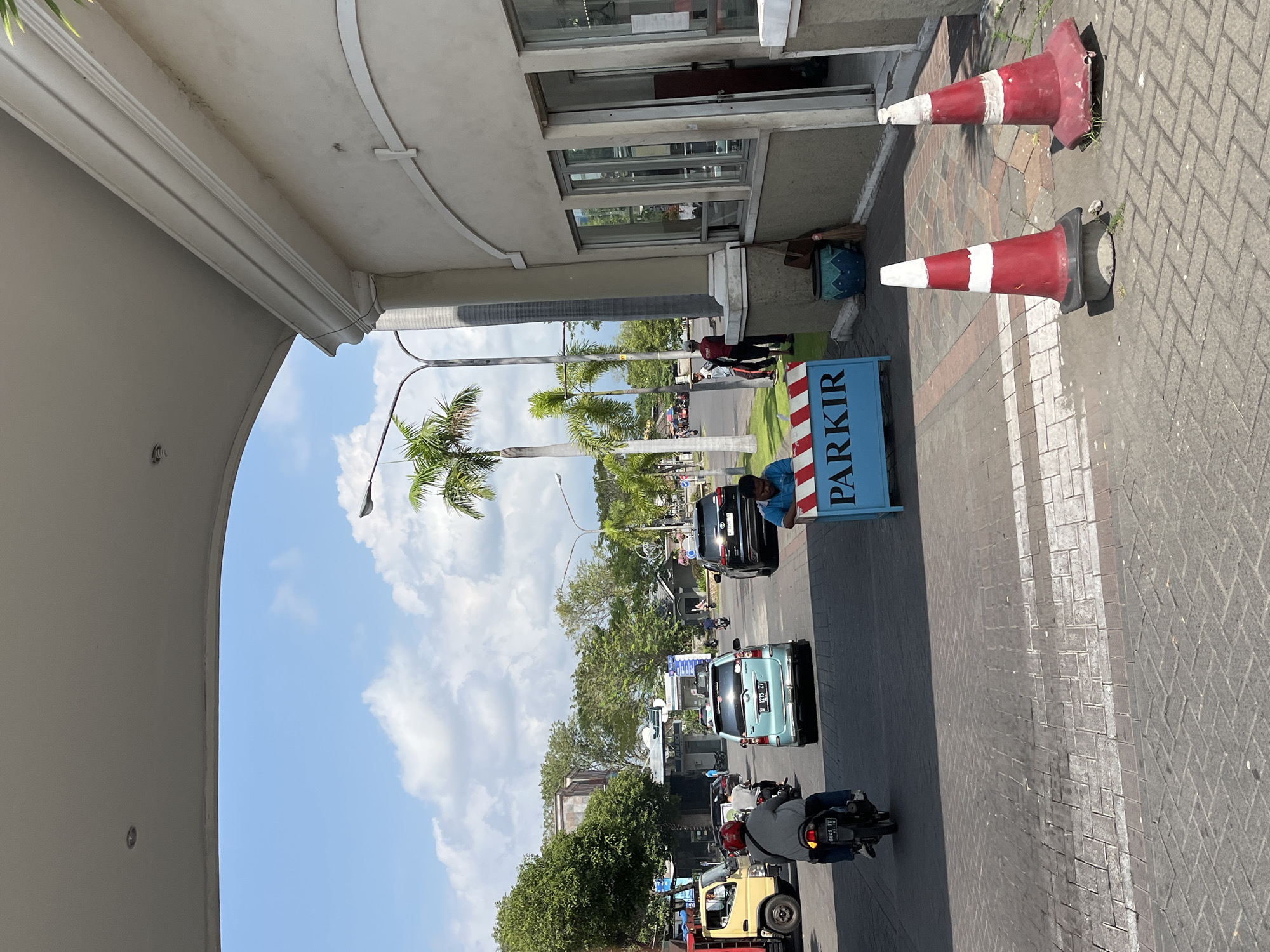 This is a ground-level, eye-level, full shot image of an entrance to what appears to be a building complex in Boro, Indonesia. The entrance is covered by a wide, white, arching structure. On the right side of the image, there is a beige building with large glass windows and an open doorway. In front of the building, there are two red and white traffic cones. A blue sign with white lettering that reads "PARKIR" is placed on the ground between the cones.
In the background, there is a street with several vehicles, including a yellow truck on the left, a light blue car, a dark SUV, and a motorcycle. There are also several palm trees and other green foliage visible. Two people are standing on the side of the street, one in a red shirt and another in a white shirt. A person is sitting on the edge of the "PARKIR" sign. A person wearing a red helmet is riding a motorcycle in the foreground, heading towards the entrance. The sky is blue with some white clouds. The ground is paved with interlocking bricks.