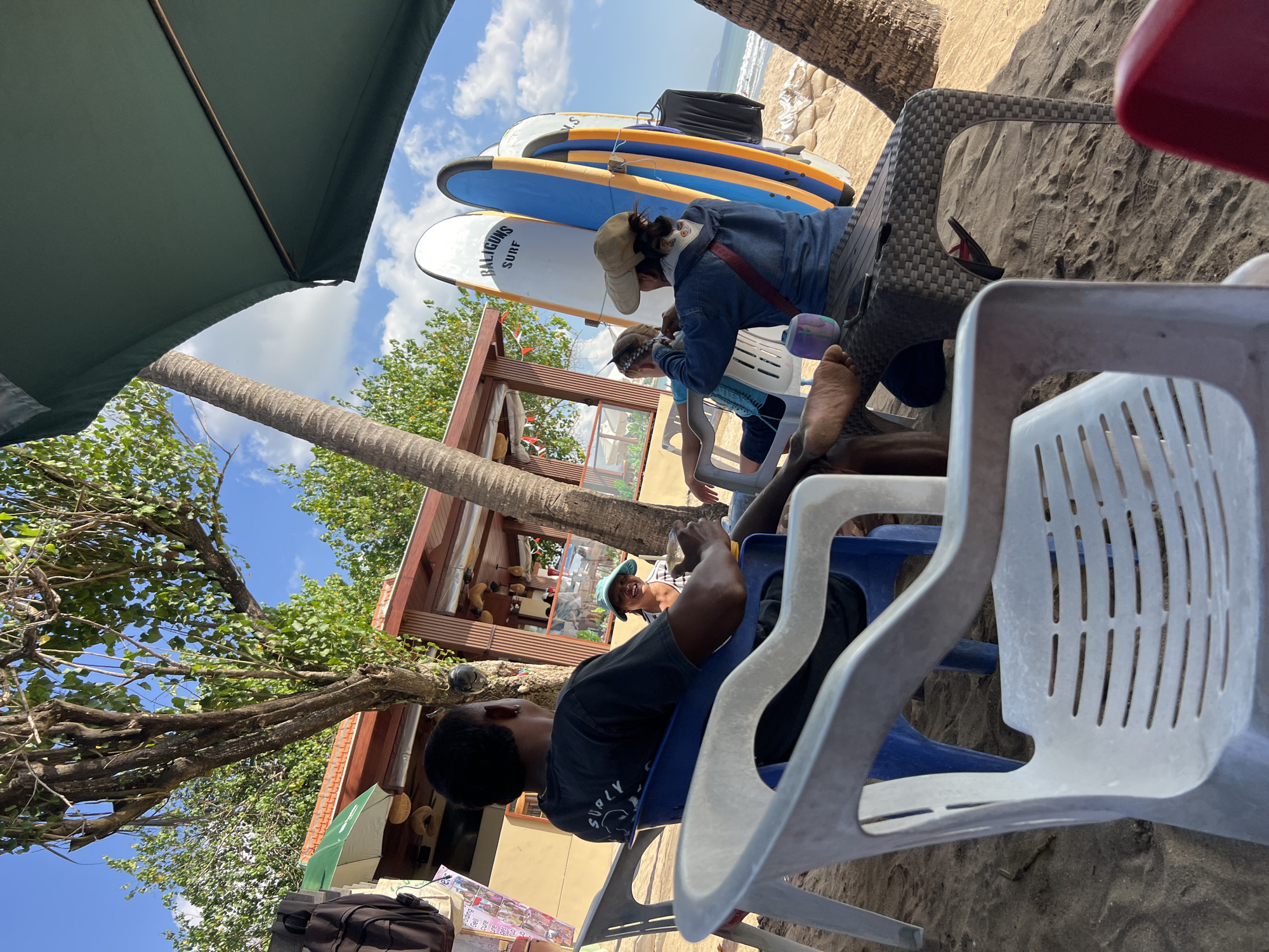 An overhead shot shows four people and surfboards on a sandy beach in Kuta, Indonesia. On the left, a man in a black t-shirt sits in a blue plastic chair, facing away from the camera. To his right, a woman in a blue denim jacket and a beige sun hat sits in a patterned chair, facing right and away from the camera. She appears to be looking at a phone or tablet. Behind her, a person in a white shirt sits in a white plastic chair, facing left and away from the camera. Between the two women, three surfboards stand upright, leaning against a building. The building has a brown roof and a beige facade, with open-air windows and a counter. Palm trees and foliage are visible in the background, along with a bright blue sky and white clouds. A green umbrella shades part of the scene. The beach is sandy, with footprints visible. The overall impression is a casual beachside setting.