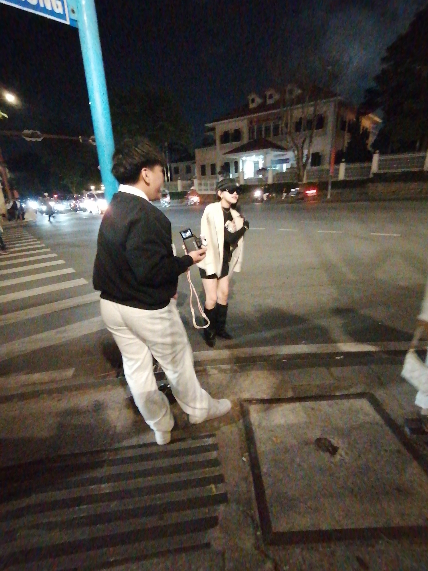 The image captures two individuals at night on a city street in Da Lat, Vietnam. The scene is illuminated by streetlights and the headlights of passing vehicles, suggesting it is after dark. The ground is wet, indicating recent rain or damp conditions.
In the foreground, on the left, a person wearing white pants and a black jacket stands facing away from the camera, looking to their right. They are holding a small electronic device, possibly a camera or phone, in their right hand, connected by a wire.
To the right and slightly behind the first person, another individual, dressed in a cream-colored blazer, black top, shorts, and knee-high black boots, is also looking to their right. They are wearing a black cap and sunglasses, obscuring their face.
A pedestrian crossing with white stripes is visible on the left side of the frame. In the background, a large building with multiple windows and a covered entrance is situated across the street. The presence of vehicles and streetlights suggests an urban environment.
A blue pole with some visible text on a sign above it is on the far left of the image. The overall mood appears casual, with the individuals seemingly engaged in conversation or observing something in the distance.