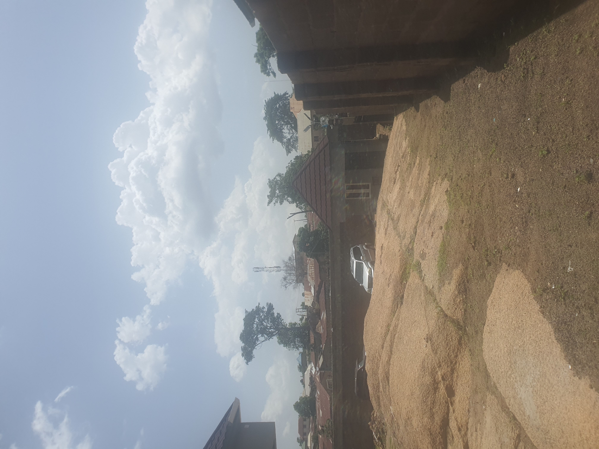 The image depicts an outdoor scene in Jos, Nigeria. The photograph is oriented vertically, with the sky and clouds dominating the left side of the frame. Puffy white cumulus clouds are interspersed with a clear blue sky, suggesting fair weather.
The right side of the image is an elevated, rocky, and sparsely vegetated terrain with a reddish-brown hue. This area appears to be a natural embankment or formation. Below this terrain, a more developed area with buildings and trees is visible. Several structures with red tiled roofs are present, indicating a residential or mixed-use area. Some trees with dense foliage are scattered among the buildings. A tall telecommunications tower can be seen in the mid-ground.
A white car is parked on a dirt path near one of the buildings. The overall impression is that of a rural or semi-urban settlement with a natural landscape feature. The lighting suggests it is daytime, likely mid-morning or afternoon, given the brightness of the sky and the shadows cast. There is no discernible text in the image. The scene appears to be peaceful and uneventful, capturing a typical day in the area.