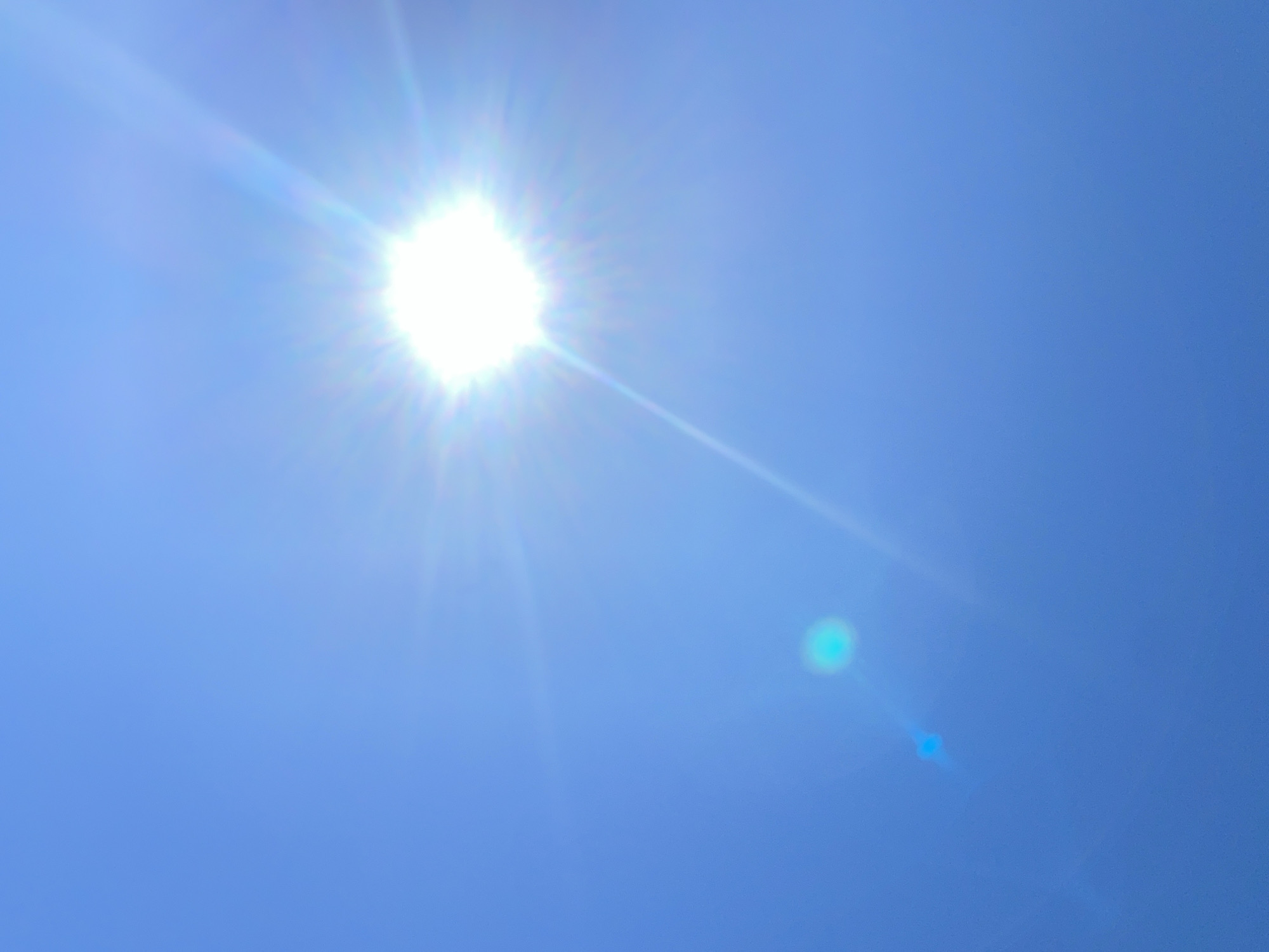 The image displays a clear blue sky with the sun prominently positioned in the upper right quadrant. The sun is very bright, emitting rays of light that extend outwards. There is a lens flare, appearing as a circular, cyan-colored spot, in the lower left quadrant of the image. A fainter, elongated lens flare is also visible near the cyan spot. The image appears to be taken outdoors on a sunny day. No specific location in the city of Police, Poland, can be determined from the image content.