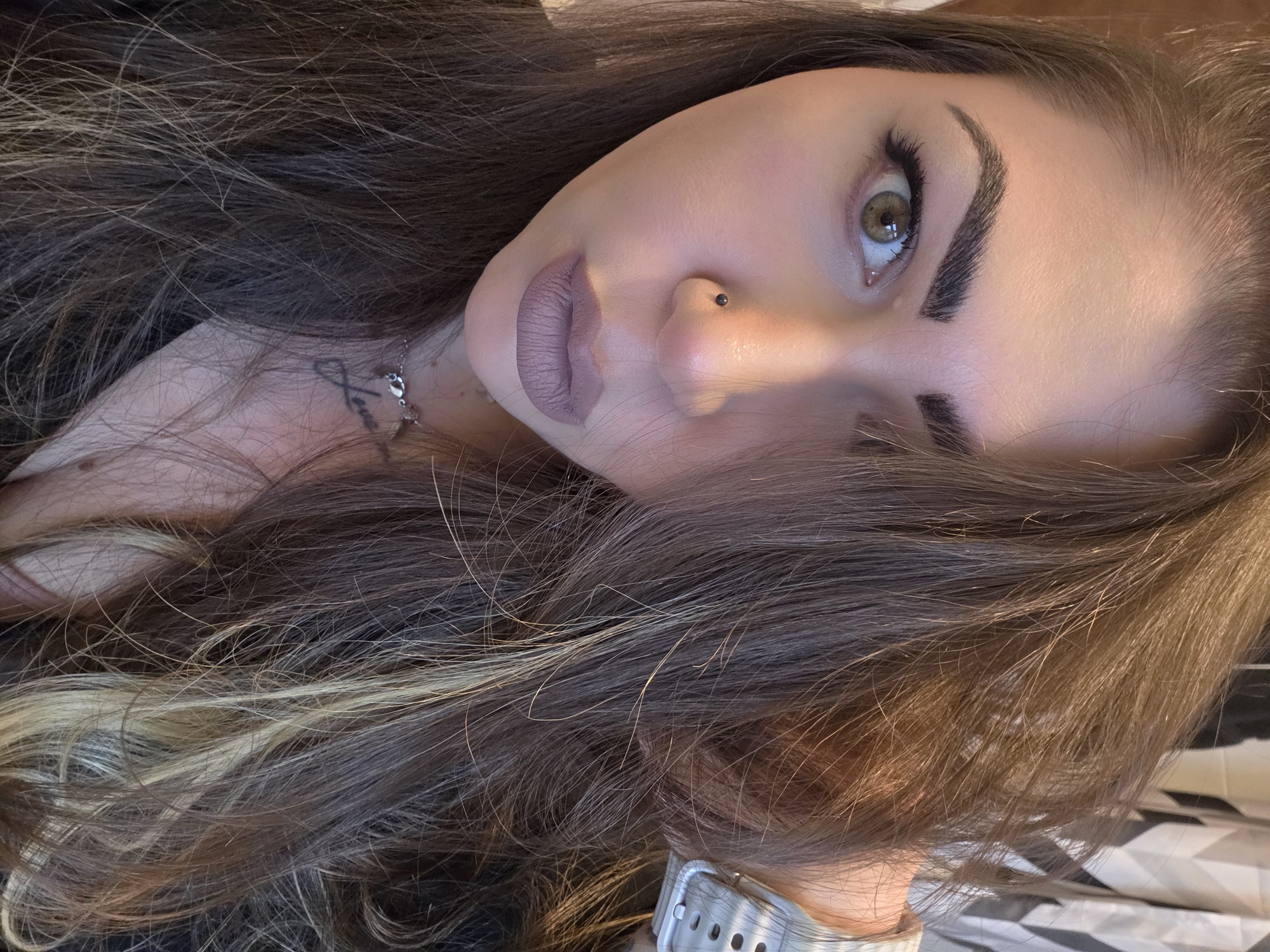 A close-up, angled portrait of a light-skinned woman with brown hair, lying down. Her hair is highlighted with blonde streaks. She has light brown eyes, prominent eyebrows, and a piercing in her left nostril. She wears mauve lipstick and appears to have eyeliner and mascara on. A thin silver chain necklace with a charm is visible on her neck, and a silver watch is on her wrist. The background is blurred and partially visible, showing a black and white geometric pattern. The location is undetermined; there is no identifiable information to ascertain its whereabouts.
