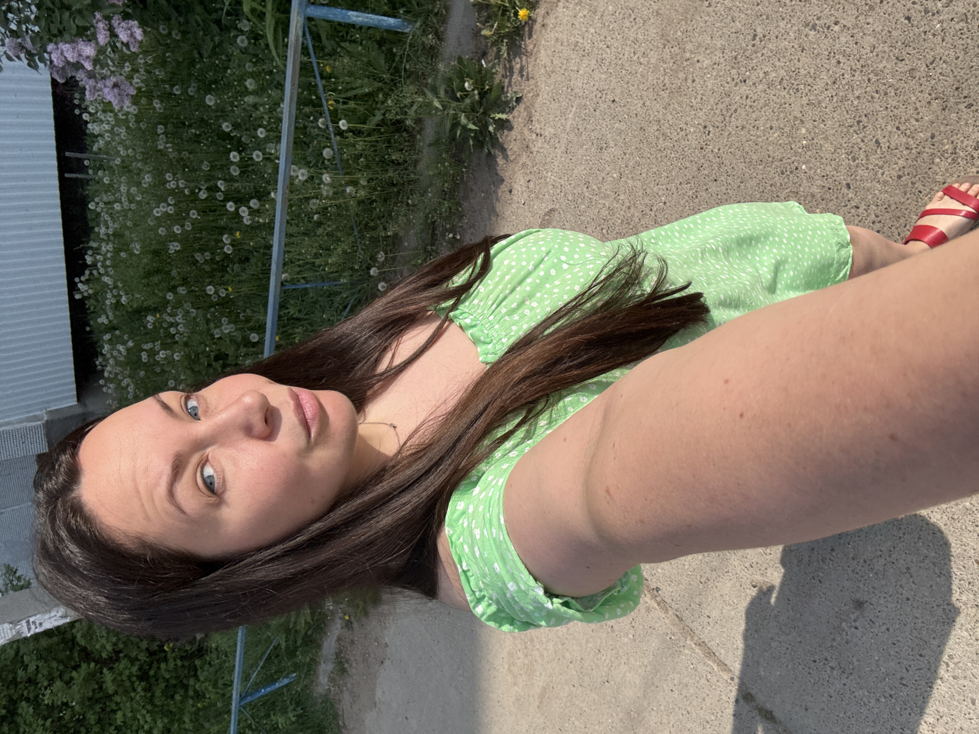 A woman with fair skin, blue eyes, and long brown hair is taking a selfie. She is wearing a green dress with white polka dots. Her left foot is visible, adorned with a red sandal. The woman is standing on a concrete surface, and in the background, there is greenery, including wildflowers and a blue metal railing. A building is also visible in the background. The location is most likely in an urban or suburban area of Chaikovsky, Russia.