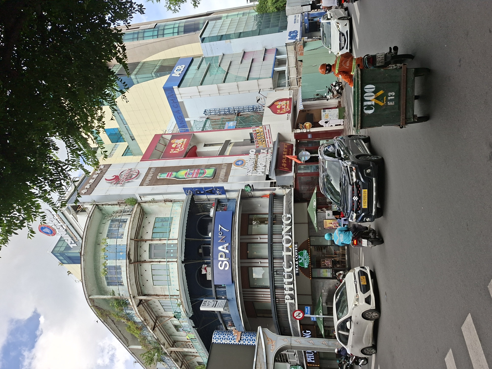 This is a street-level, wide-angle photograph taken in Thủ Đức, Vietnam. The image is vertically oriented and appears to be taken at eye-level, with the street dominating the lower half of the frame and buildings lining the right side. A lush green tree with dense foliage partially obscures the upper left portion of the image.
The scene depicts a busy street with several vehicles, including cars and motorcycles. In the foreground, a person wearing an orange uniform and helmet is near a green dumpster, suggesting a sanitation worker. A white car is parked on the right side of the street, and a black car is parked further down. A motorcycle with a rider in a blue uniform is also visible. The buildings are multi-story structures with various businesses indicated by signage. Notable text includes "ACB" (likely a bank), "SPA N°7," "PHUC LONG TEA & COFFEE," "TSINGTAO," and "SRI RAJA POJONAL RESTAURANT." The architecture suggests a mix of modern and older styles, with some buildings featuring balconies and balconies with greenery.
The time of day appears to be daytime, as indicated by the bright sky and sunlight casting shadows. The weather seems clear, with blue sky and white clouds visible. The overall impression is one of an active urban environment with commercial activity and traffic. The presence of street vendors, signs in Vietnamese, and the vehicle license plates are strong indicators of the location in Vietnam.
