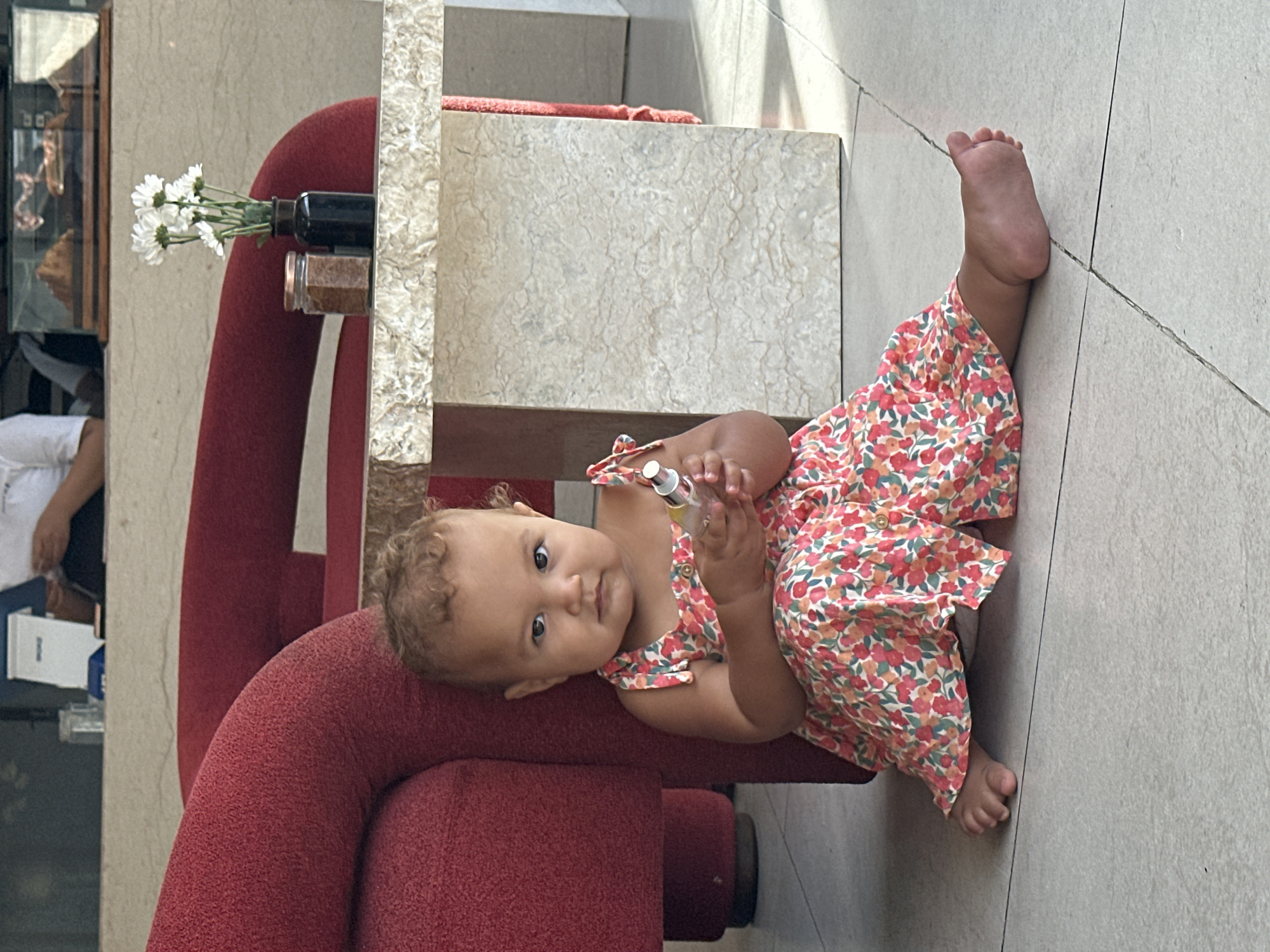 A young female child with light brown curly hair and brown eyes sits on a tiled floor, wearing a floral-print dress. She is holding a clear cylindrical object with a silver top in her right hand. Behind her, a plush red armchair is partially visible, and a marble-topped table stands with a dark vase containing white flowers and a small jar of spices. In the background, a glass display case exhibits pastries, and a person in a white shirt is seated at a counter. The scene appears to be a cafe or hotel lobby. This location is not identifiable as being in the city of Mengwi, Indonesia.