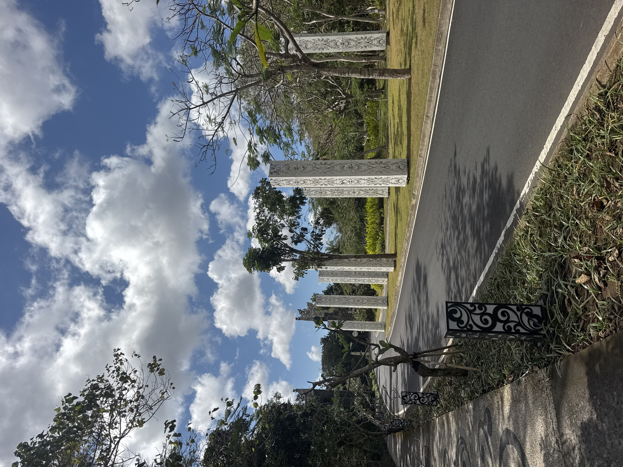 This is an outdoor, eye-level, vertical photograph of a road in Kuta Selatan, Indonesia, featuring decorative pillars. The road is paved with asphalt and bordered by a concrete curb and a white stripe. To the left of the road is a paved walkway with a textured surface, shaded by trees. Along the walkway, there are black decorative lamps with intricate swirling patterns. Beyond the walkway and to the left of the road is a dense line of trees.
The road itself is lined with a manicured lawn, where several tall, white, rectangular pillars stand at intervals. These pillars are adorned with detailed, ornate carvings in a light color, creating a striking visual contrast against the white surface and the green grass. Some of the pillars are partially obscured by trees. Behind the pillars and lawn, there is a background of more trees and vegetation, with some taller structures or gates visible in the distance.
The sky above is a vibrant blue, dotted with fluffy white cumulus clouds. The lighting suggests it is daytime, with shadows cast by the trees on the road and walkway. The overall scene depicts an entrance or pathway to a well-maintained area, likely a resort, park, or cultural site, with a tropical ambiance due to the lush greenery and warm climate suggested by the clear sky. No people or animals are visible in the frame.