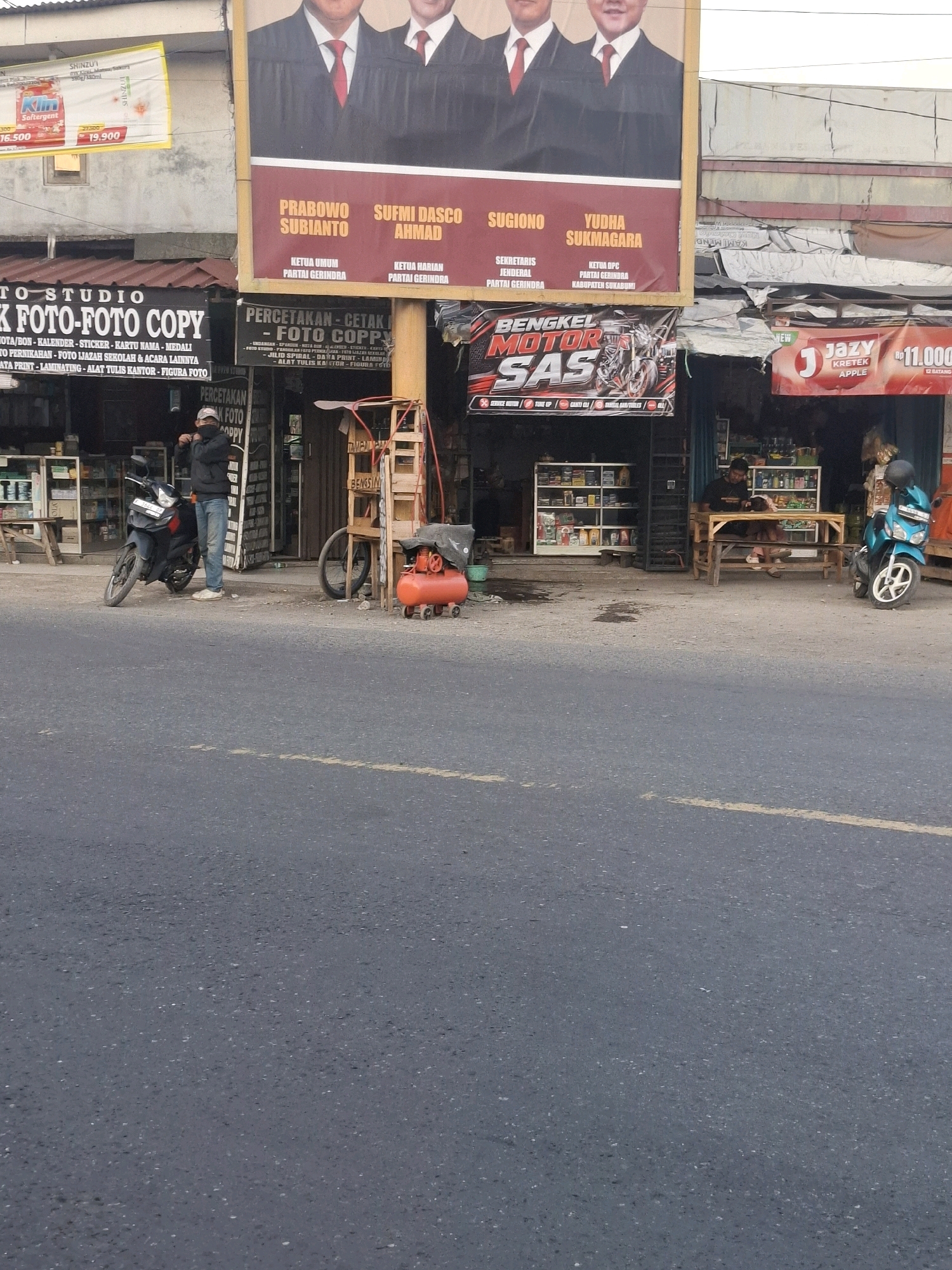 This is a street-level photograph taken in Sukaraja, Indonesia, capturing a commercial area. In the foreground, a paved road with yellow lane markings is visible. On the right side of the road, a bright blue scooter is parked. Further back, there is a roadside stall with a wooden table and benches, where two individuals are seated, one appearing to be reading. Adjacent to this is a motorcycle repair shop with a banner displaying "BENGKEL MOTOR SAS." To the left of the repair shop is a photo studio and copy center with a sign that reads "TO-STUDIO FOTO-FOTO COPY." A man wearing a black jacket and blue jeans stands beside a black motorcycle in front of this establishment. Dominating the upper portion of the image is a large billboard featuring portraits of four men and political text. The buildings behind the shops are simple, one-story structures. An orange air compressor is positioned on the sidewalk near the repair shop.