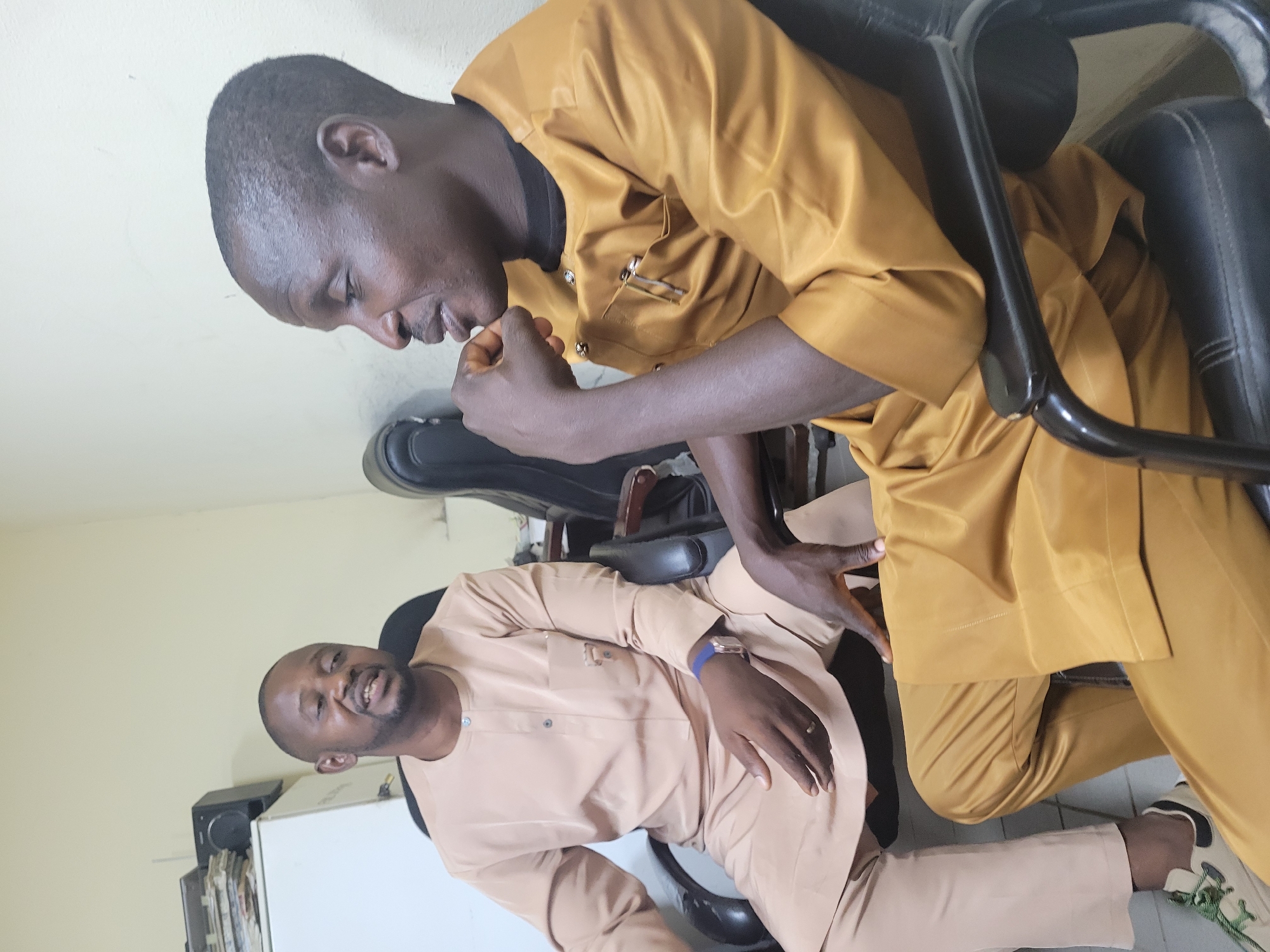 Two men are seated in office chairs, facing each other. The man on the left is wearing a light brown kaftan and pants, with a watch on his left wrist. He is speaking with his mouth open. The man on the right is wearing a golden-yellow kaftan and pants. He has his right hand resting on his chin, and a pen clipped to his left breast pocket. The background appears to be an office setting in Abuja, Nigeria, with a filing cabinet visible to the left.