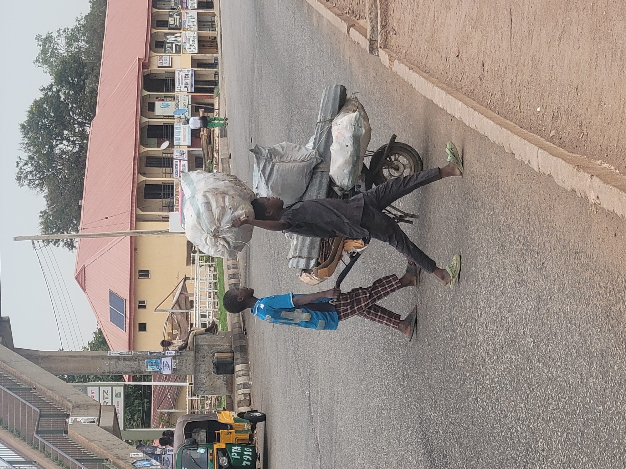 Two young men are struggling to push a hand truck overloaded with large sacks down a paved street in Gura Top, Nigeria. One man, wearing a black shirt and dark pants, is pushing the load from behind while the other, wearing a blue shirt and checkered pants, is pulling from the front. The hand truck is loaded with at least four large white sacks. A yellow and green "keke" (tricycle taxi) is parked nearby. Buildings line the street, including one with a red roof and solar panels. Power lines and a pedestrian bridge are also visible.