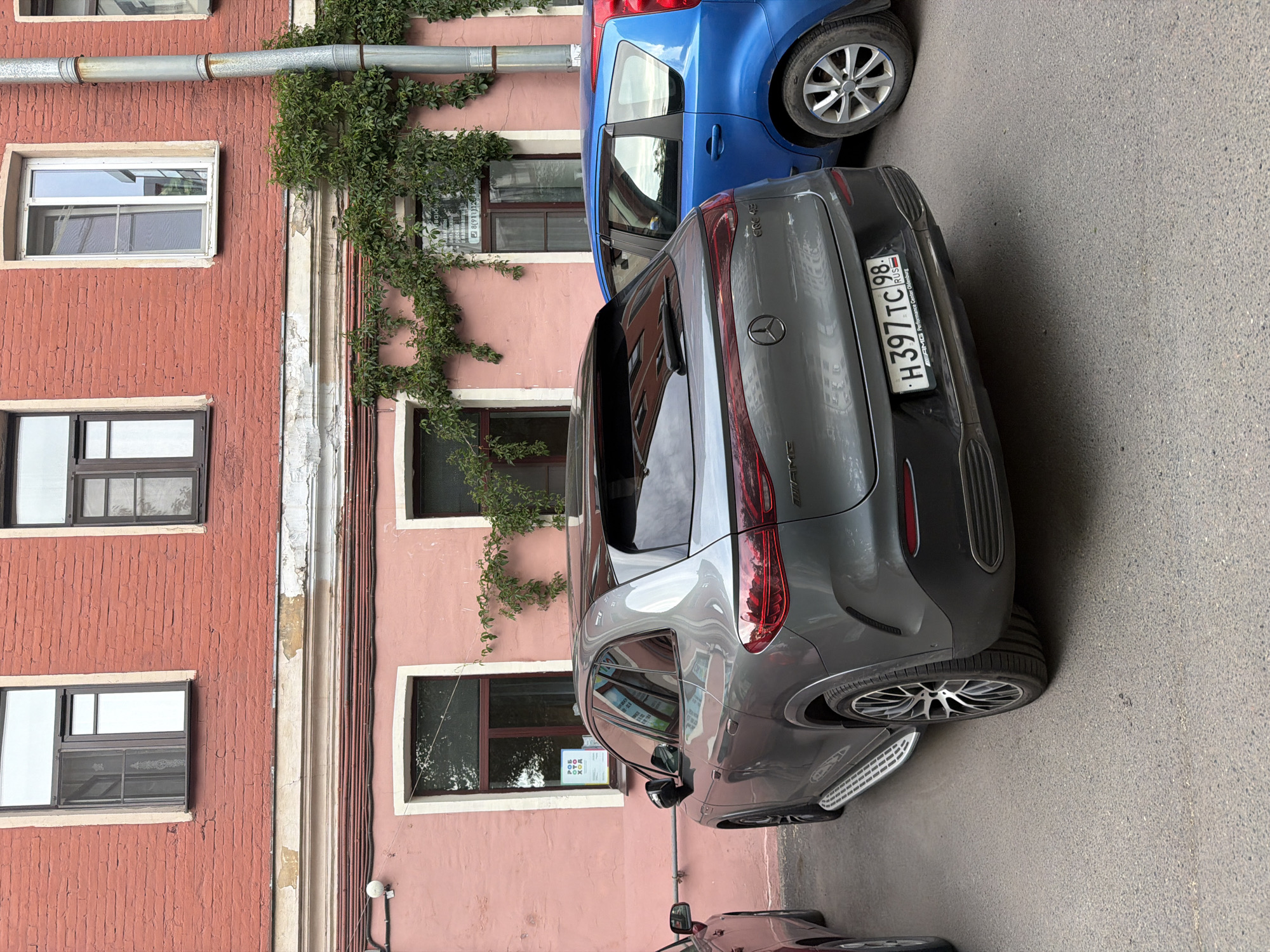 The media file shows two cars parked on a street next to a pink and red building. The building has several windows and is adorned with ivy growing on its facade. The cars are a blue hatchback and a grey Mercedes-Benz SUV. The license plate of the grey car is visible and reads "H397 TC 98". The scene appears to be outdoors during the daytime, with no clear indications of weather conditions. The setting is likely an urban environment given the street and building. There are no people visible in the image. The overall impression is a candid snapshot of parked vehicles on a street.