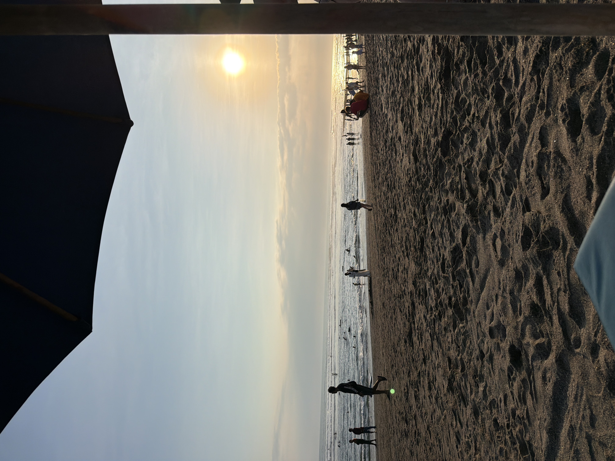 The image depicts a beach scene in North Kuta, Indonesia, during sunset. A large dark blue umbrella is in the foreground, partially obscuring the view. The beach is covered in dark sand with visible footprints. Several people are scattered across the sand and in the water. Some individuals are walking on the beach, while others are in the ocean, some appearing to be surfing. The sun is low in the sky, casting a warm glow and reflecting off the water. Clouds are present in the sky, catching the sunset colors. The overall atmosphere suggests a tranquil evening on a popular beach.