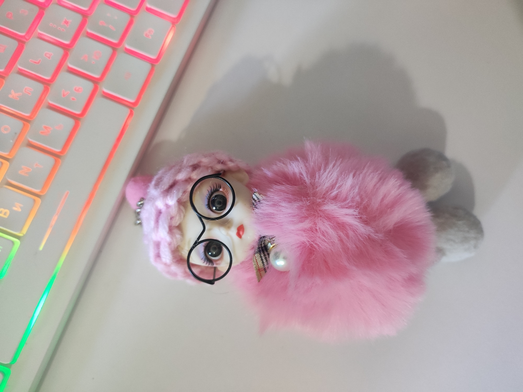 A close-up, top-down shot shows a plush toy with pink fur lying on a white surface next to a white keyboard with RGB lighting. The toy has a doll-like face with large black eyes and glasses, a small red mouth, and a pink knitted hat. It is adorned with a pearl charm and a plaid ribbon. The keyboard displays a gradient of colors from green to orange and pink on its keys. The overall scene is static and appears to be an indoor setting. There is no discernible information about the location in Cherkasy, Ukraine.