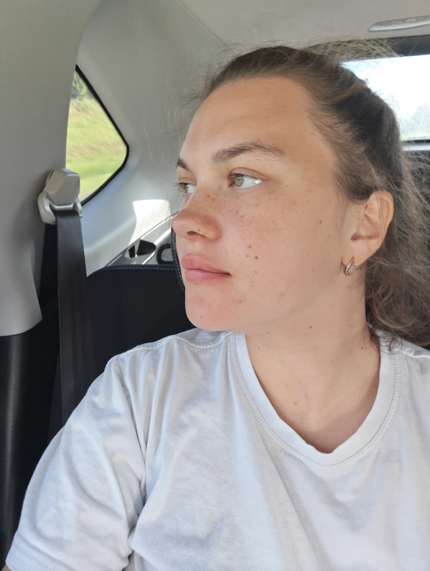 A woman with light skin and freckles is sitting in the backseat of a car. She is wearing a white t-shirt and a small earring. She is looking out the window. The car has a black seat and a grey interior. A seatbelt is visible. Through the window, green vegetation is seen.